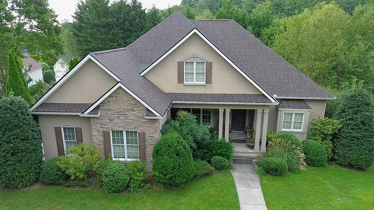 A two-story house with a gray roof, beige walls, and stone accents. The house has front steps, white columns, and an upper window with brown shutters. Surrounding the house is lush greenery and trees.