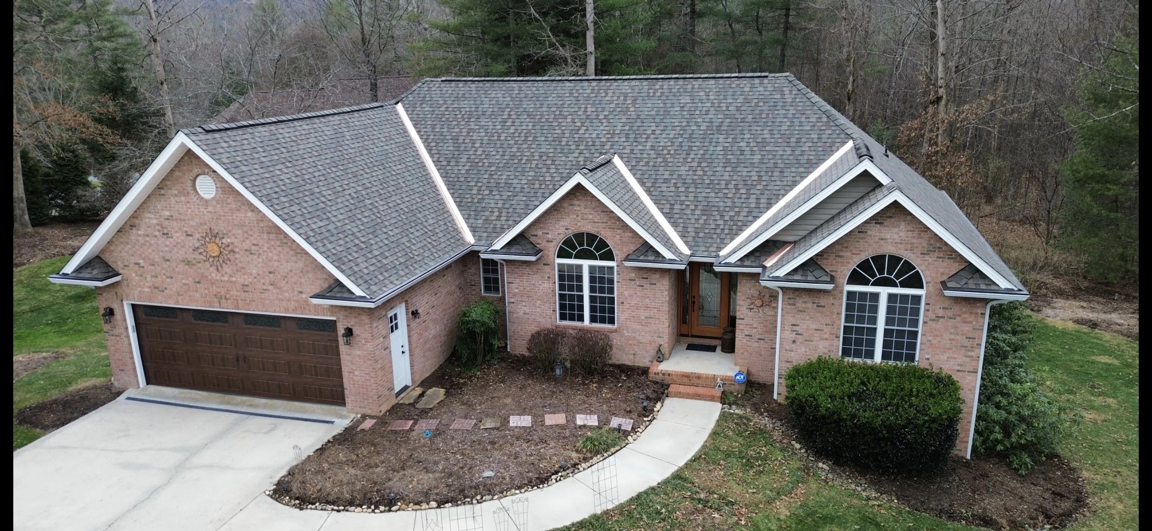 A brick house with a brown garage door, a front porch with a wooden door, and large arched windows, surrounded by trees, with a curved walkway leading to the entrance.