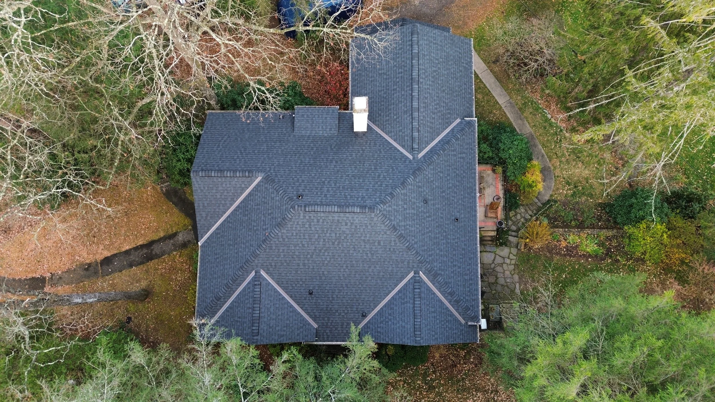 Aerial view of a house with a dark gray roof surrounded by trees with autumn foliage and a curved stone pathway leading to a backyard patio area.