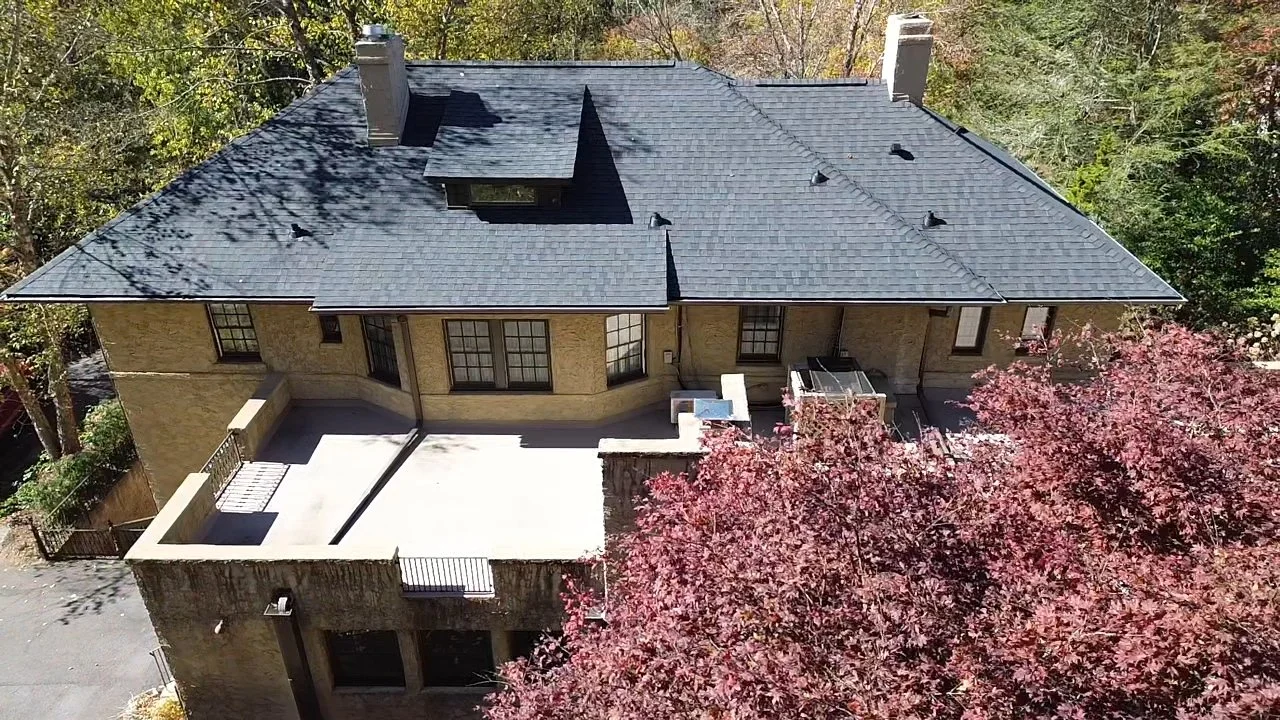 Aerial view of a house with a black tiled roof, yellow brick walls, and a concrete patio, surrounded by trees with autumn foliage.