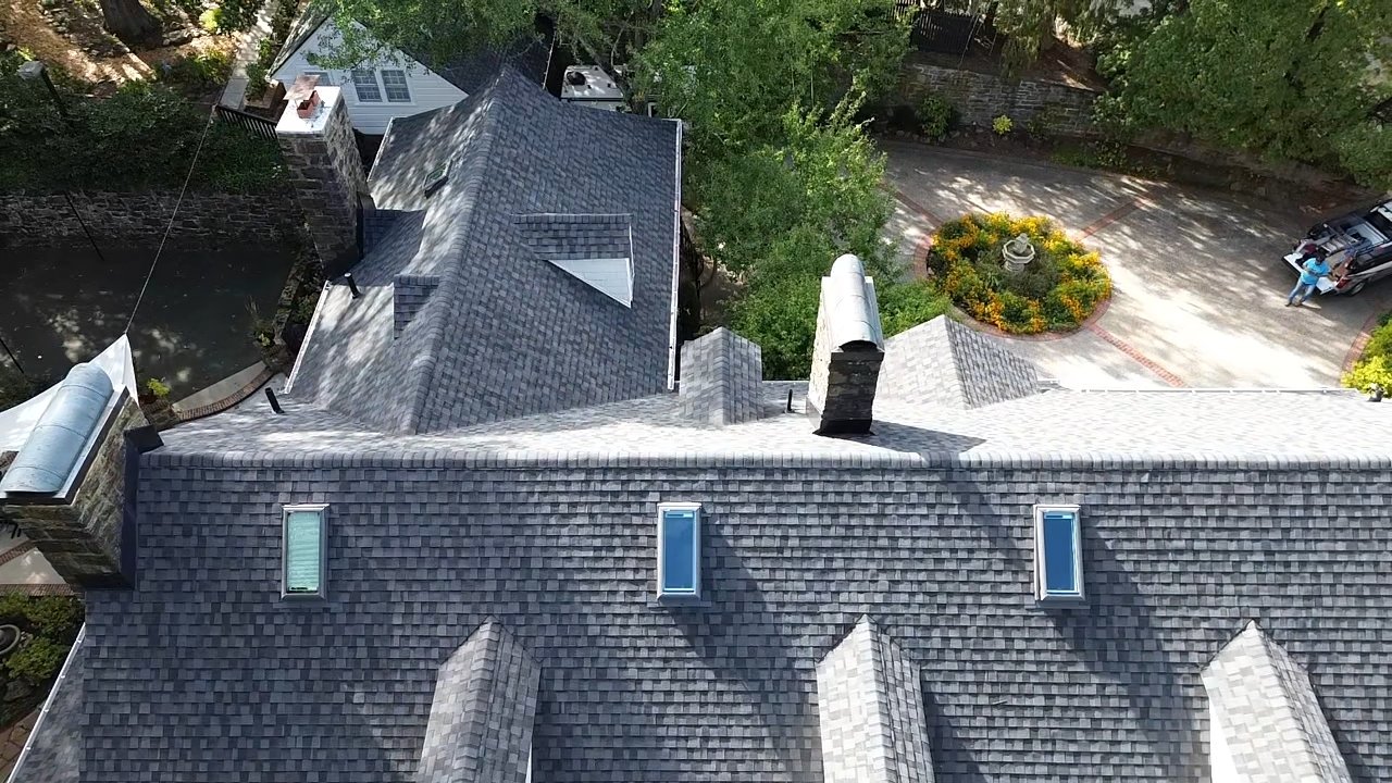 Aerial view of a townhouse with gray shingle roof, skylights, and chimneys, surrounded by trees and a parking area with a person near a car.