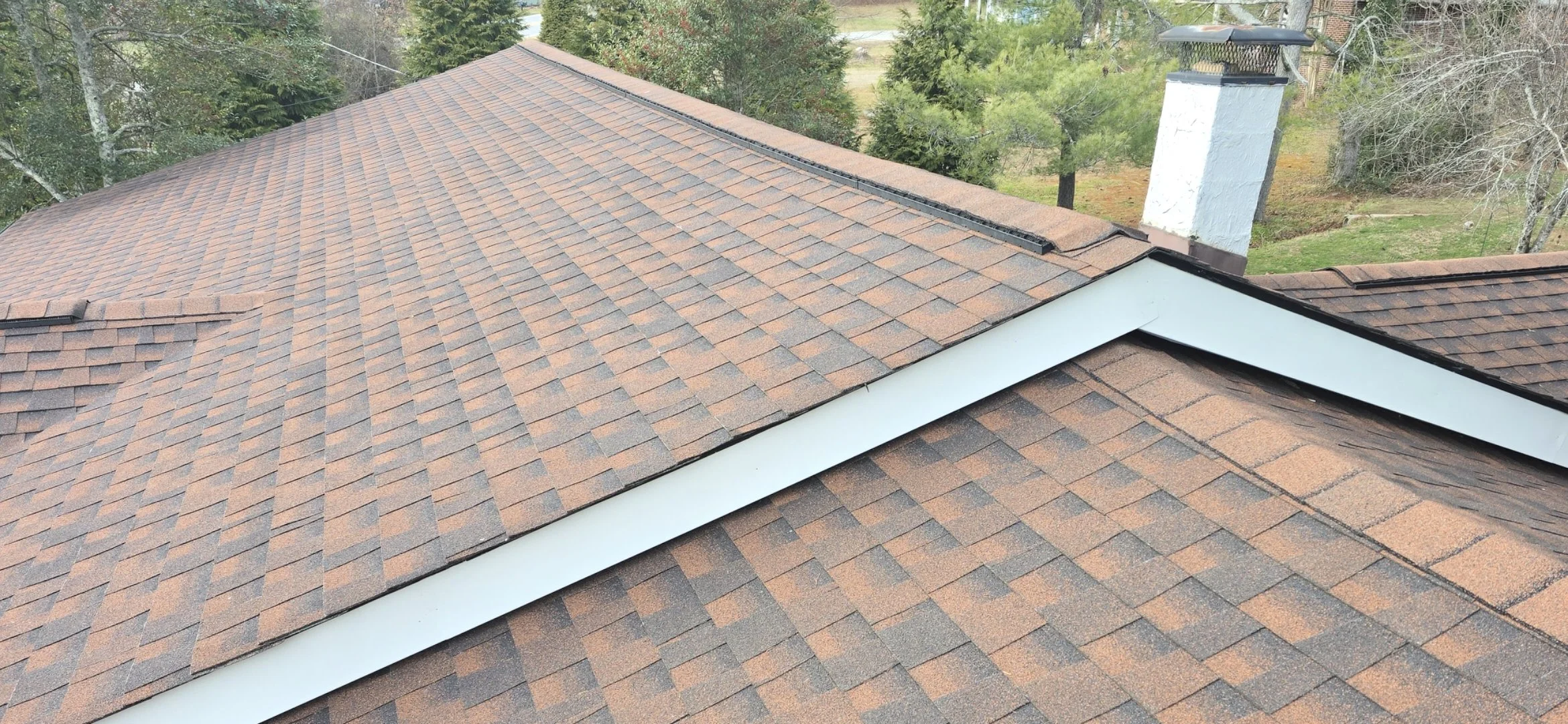 A residential roof with brown asphalt shingles and a white soffit, surrounded by trees and a yard with a chimney.