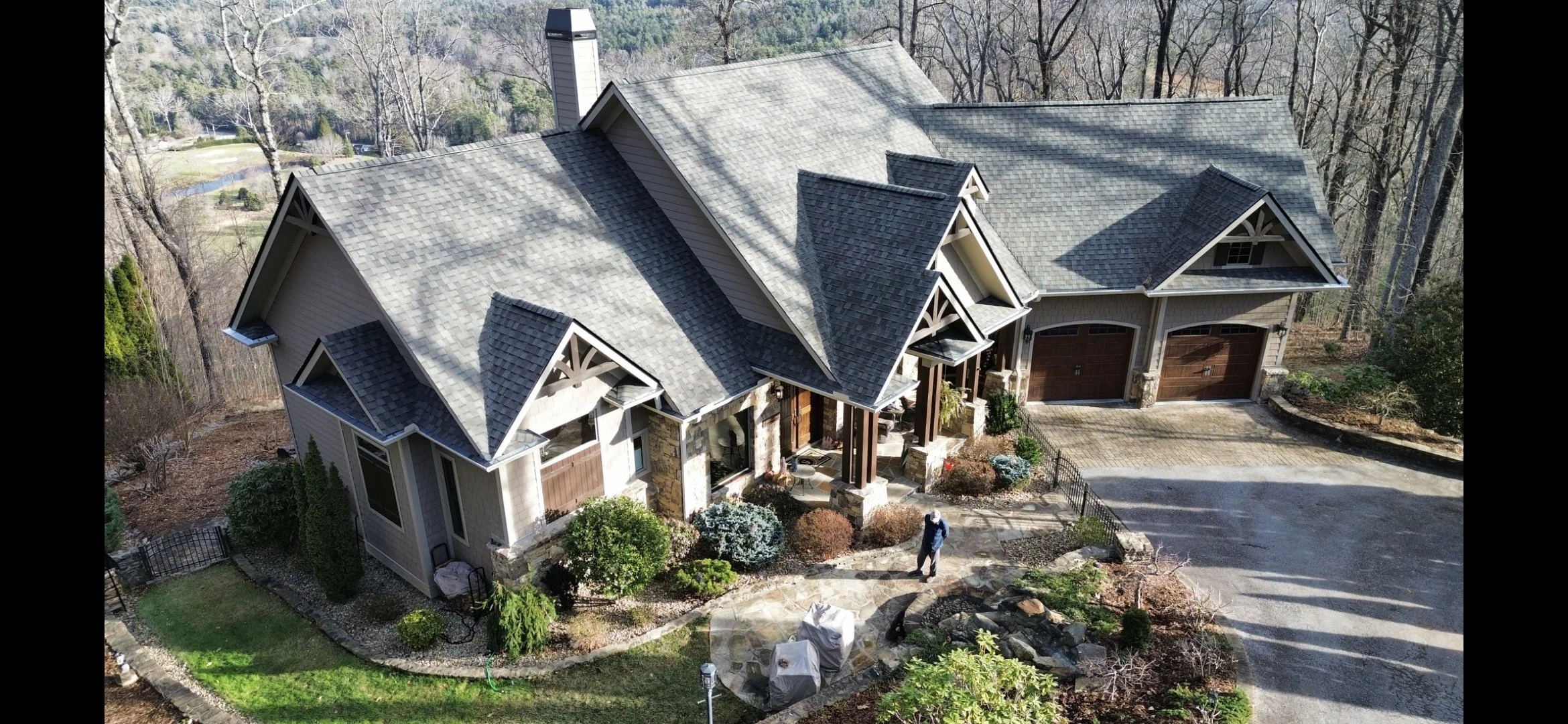 An aerial view of a large, modern house with a multi-gabled roof, two attached garages with wooden doors, stone accents, a porch, and landscaped yard with bushes and trees, surrounding a wooded area and a pond in the background.