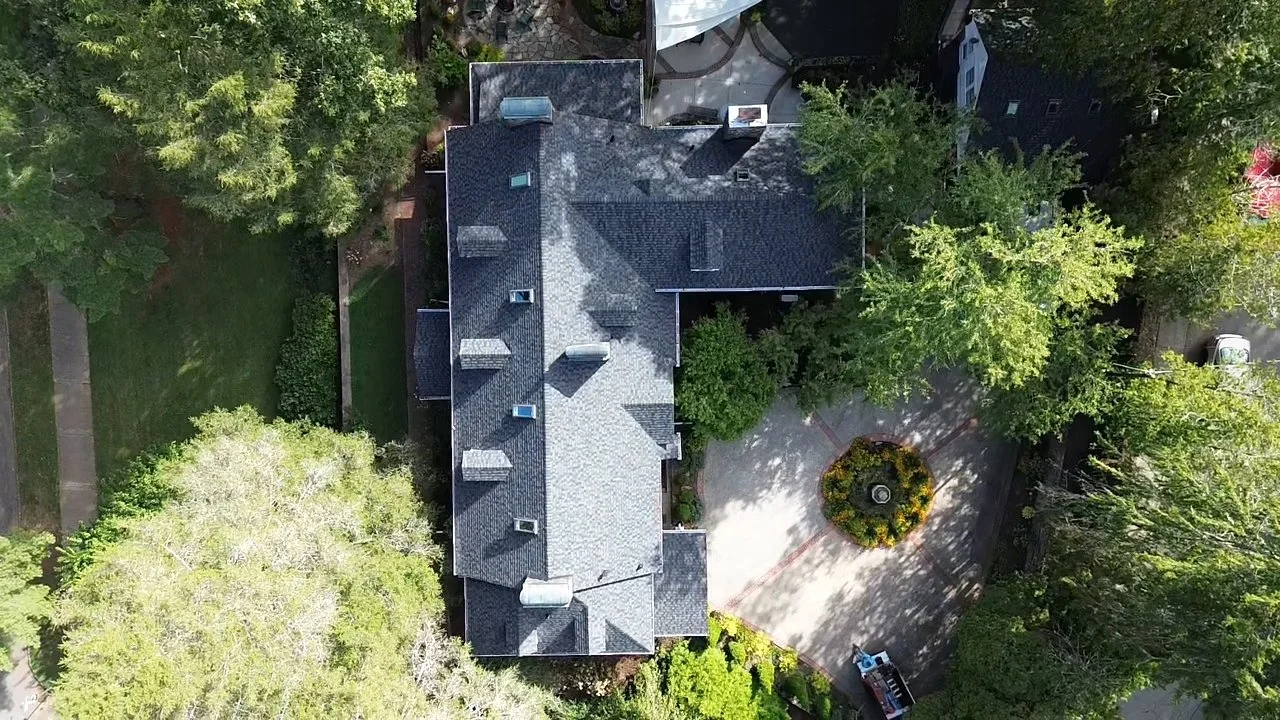 Aerial view of a house with a grey roof, driveway with a circular landscaped island, and surrounded by green trees and lawns.