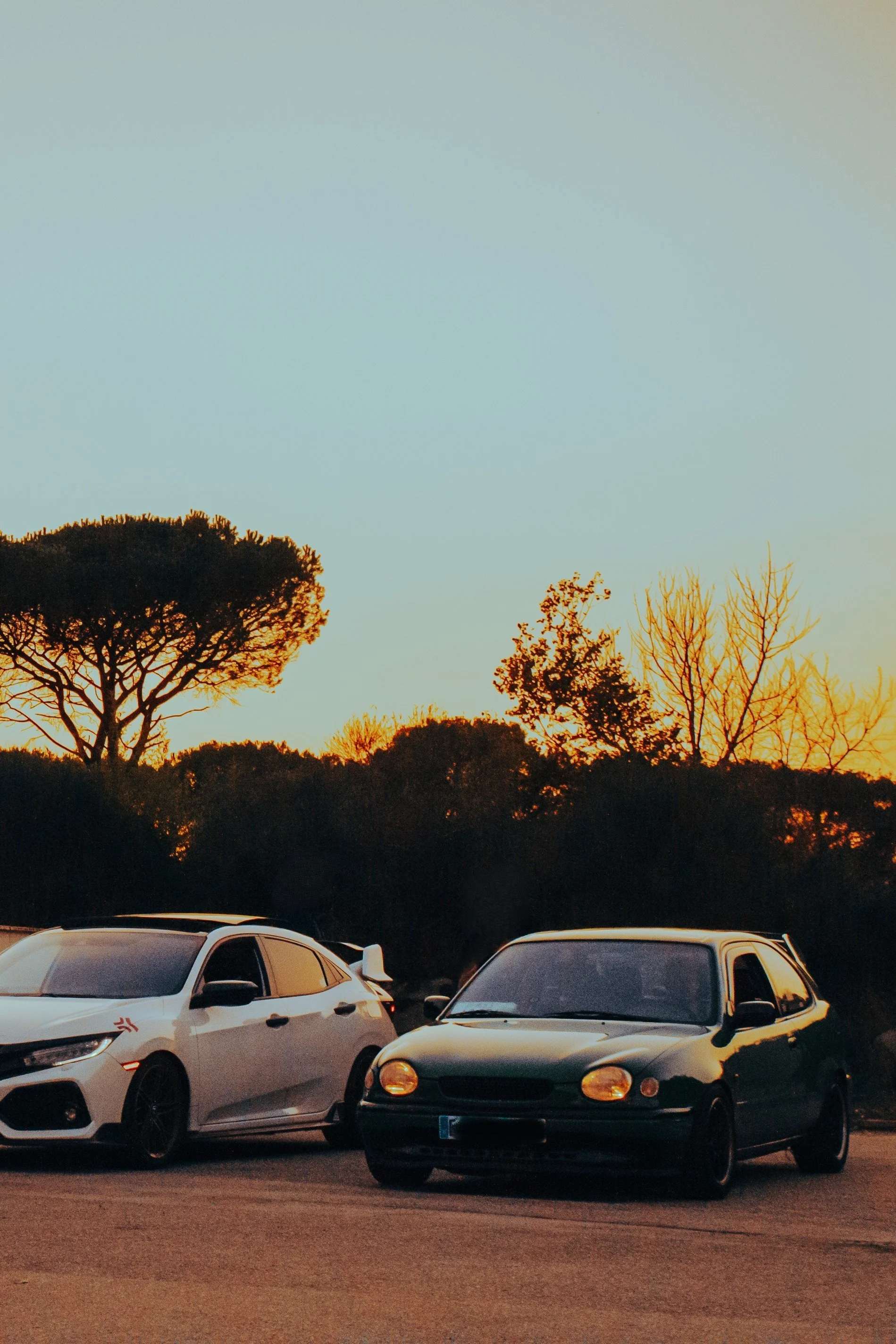 Dos autos estacionados en un área de parking bajo un cielo al atardecer, con árboles de fondo.
