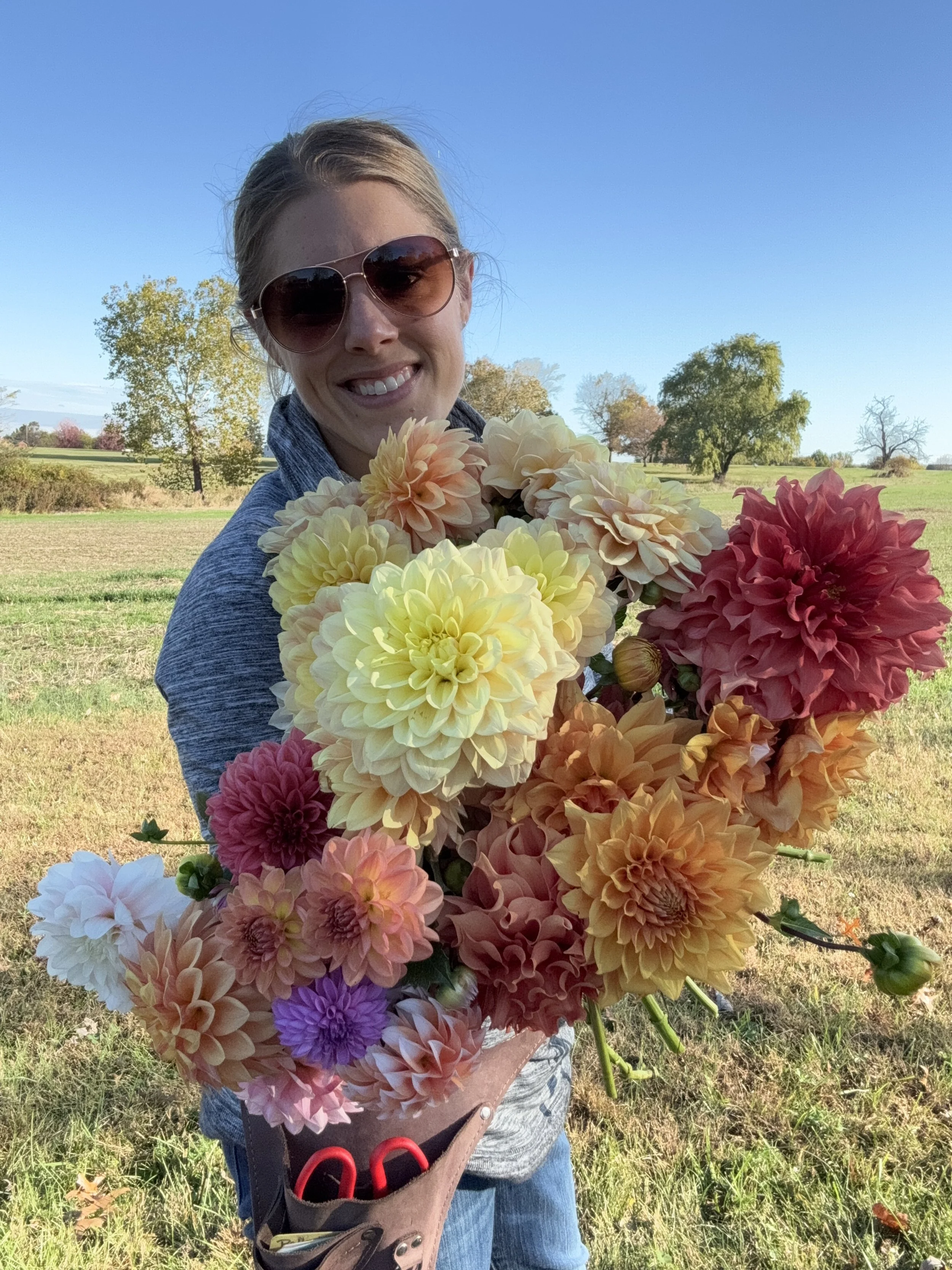 A woman holding a large bouquet of colorful flowers outdoors on a sunny day, wearing sunglasses and a blue jacket.