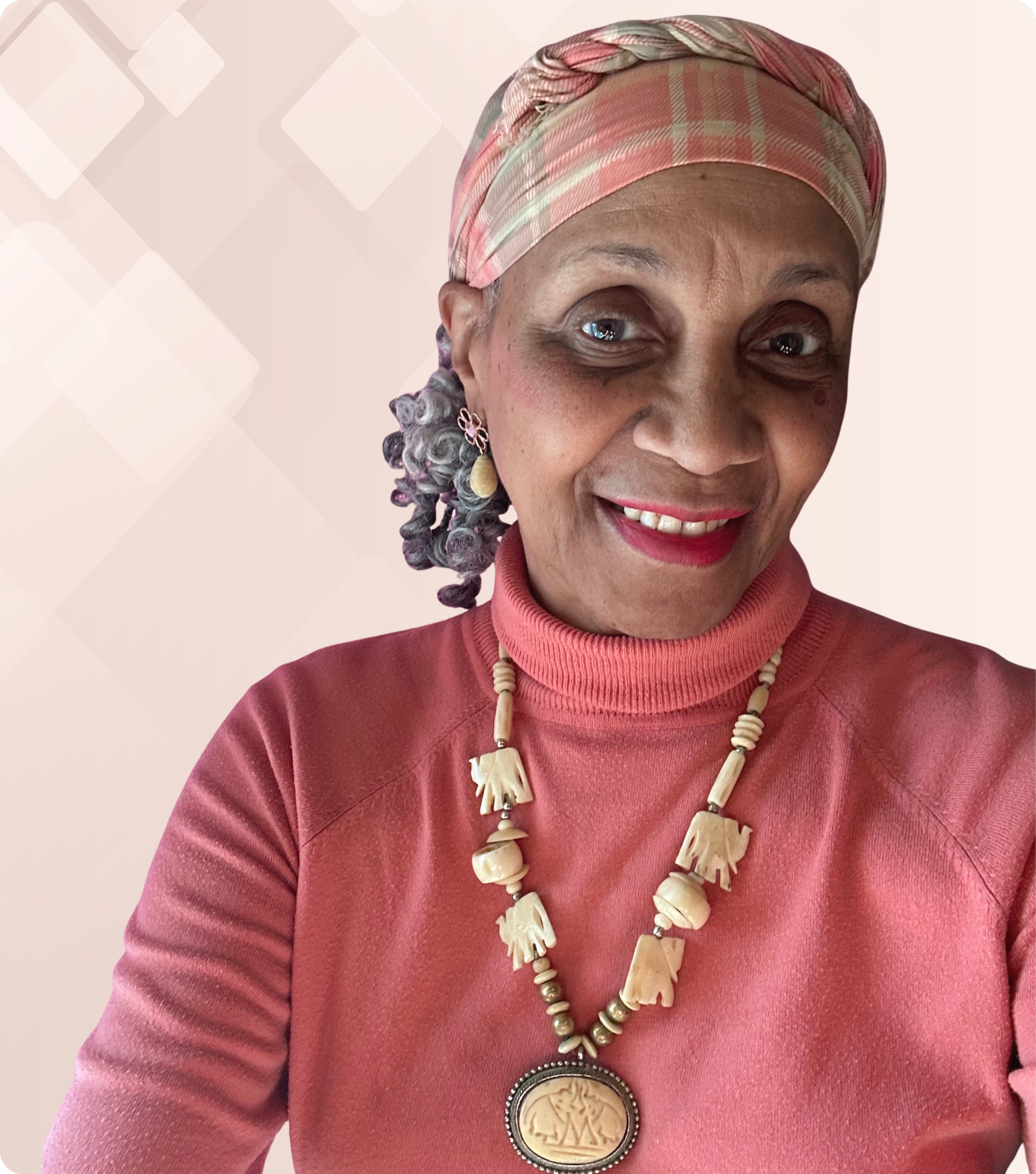 A woman smiling, wearing a pink headscarf, pink turtleneck top, and a beaded necklace with carved elephant charms and a large oval pendant featuring a tree design.