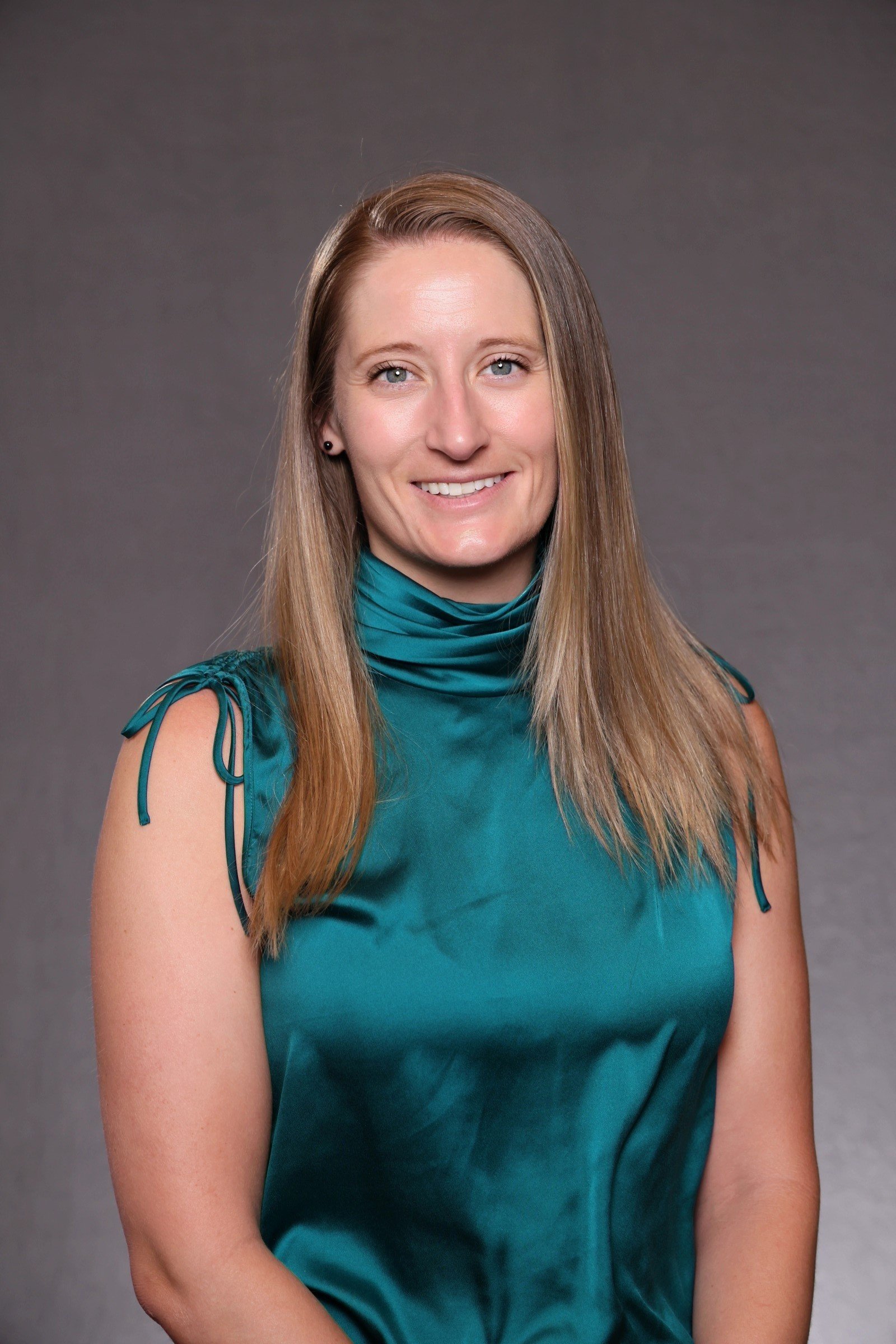 A woman with long, straight blond hair, wearing a teal satin sleeveless top with tie details on the shoulders, smiling against a plain gray background.