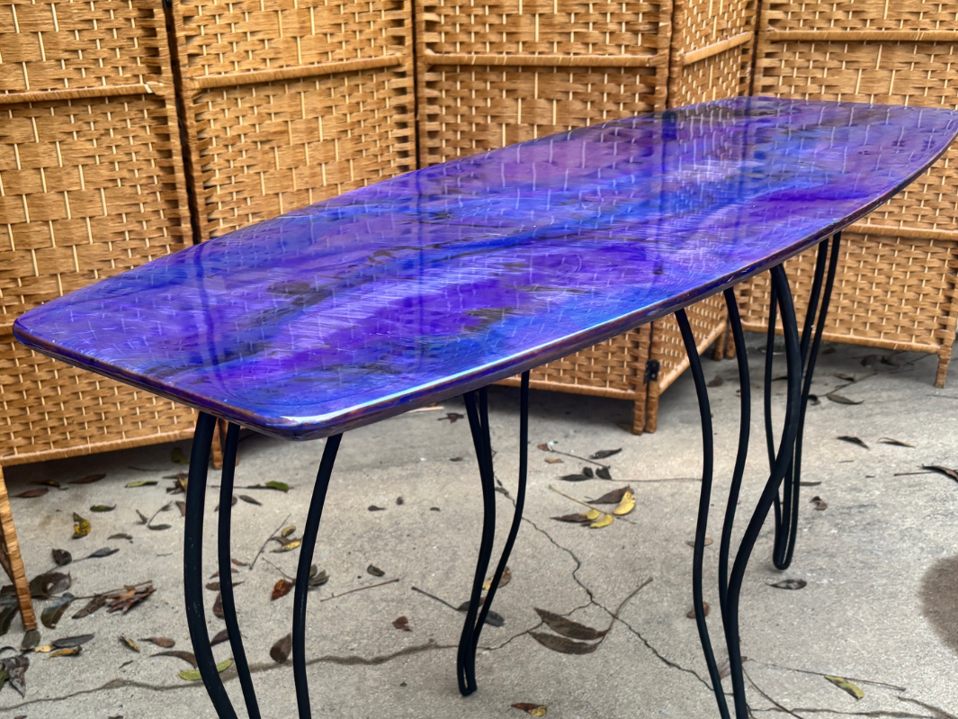 A glass-top table with a blue and purple marbled design, supported by black metal legs that are curved and intertwined. The table is outdoors, on a cracked concrete surface with scattered dry leaves. In the background, there is a woven wicker room divider.