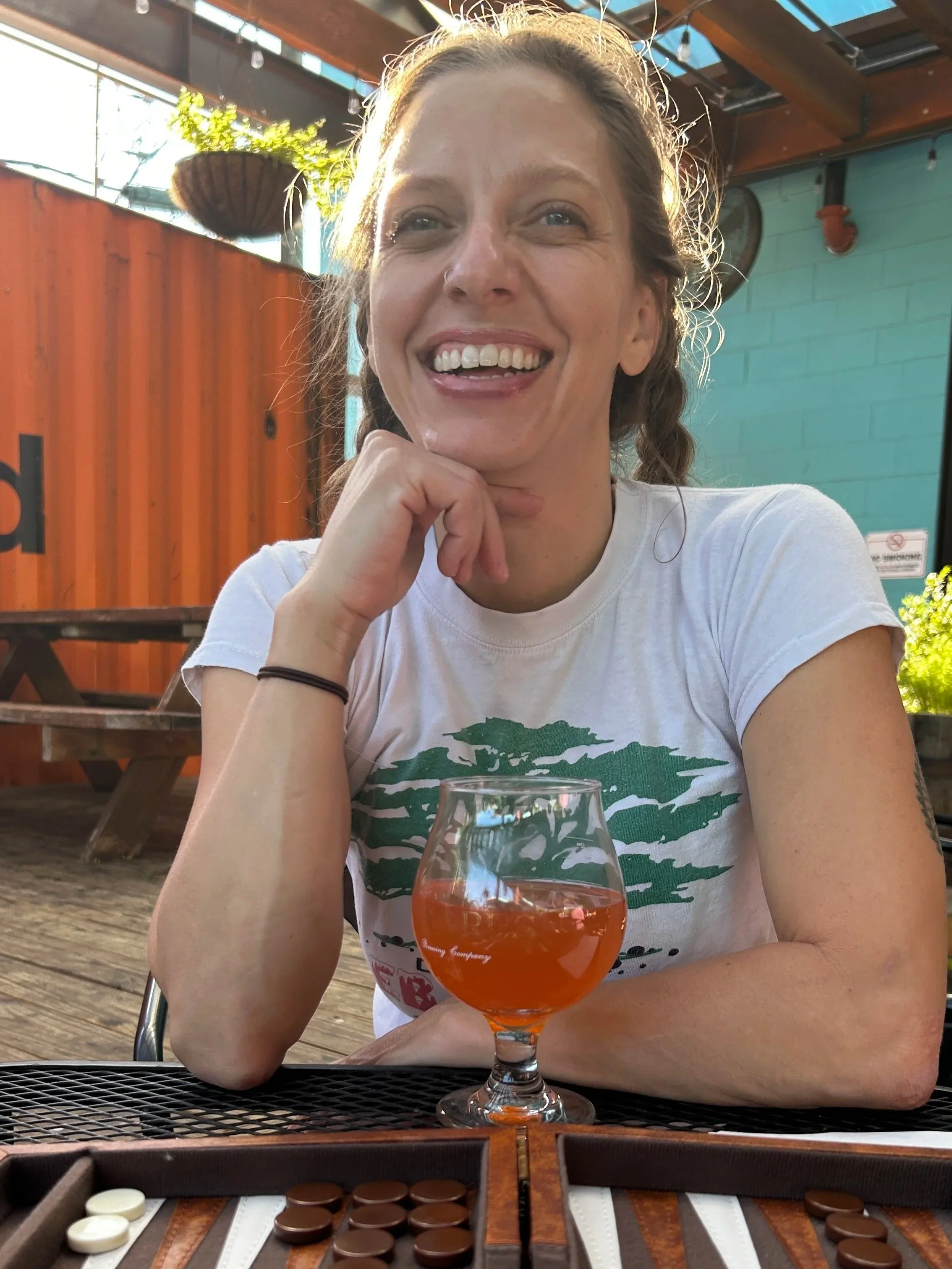 A woman with braided hair smiling and resting her chin on her hand, sitting at an outdoor table with a glass of orange-colored beverage and game pieces on the table. The background features a colorful patio with a teal wall, orange fencing, hanging planters, and a wooden structure.