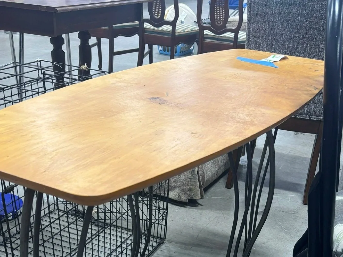 Wooden dining table with a blue label or pamphlet on it, surrounded by chairs, in a thrift store or furniture shop.