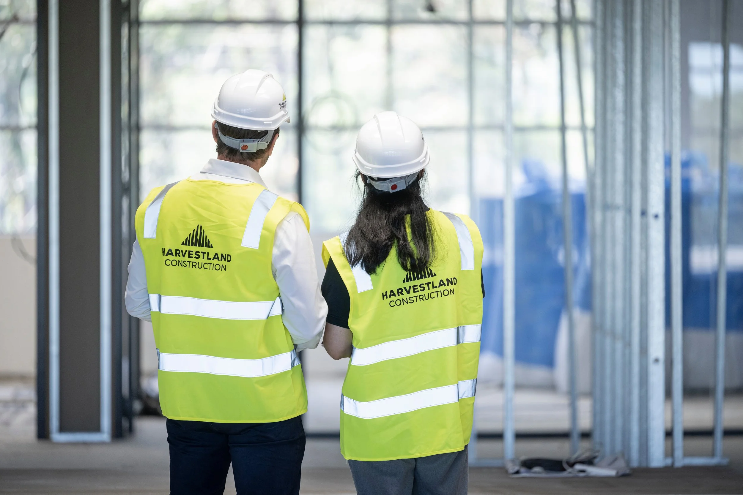 Two construction workers wearing yellow safety vests and white hard hats stand inside a building under construction, looking at the framed walls.