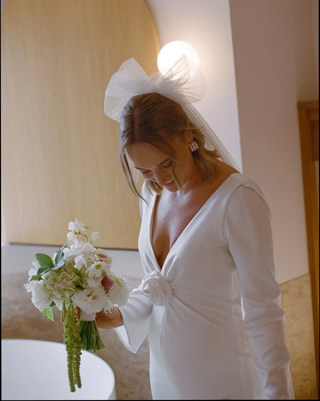 Bride holding a bouquet of white flowers, wearing a white dress with a deep V-neck and a large bow at the waist, with a veil and a large decorative bow in her hairstyle, smiling and looking down.