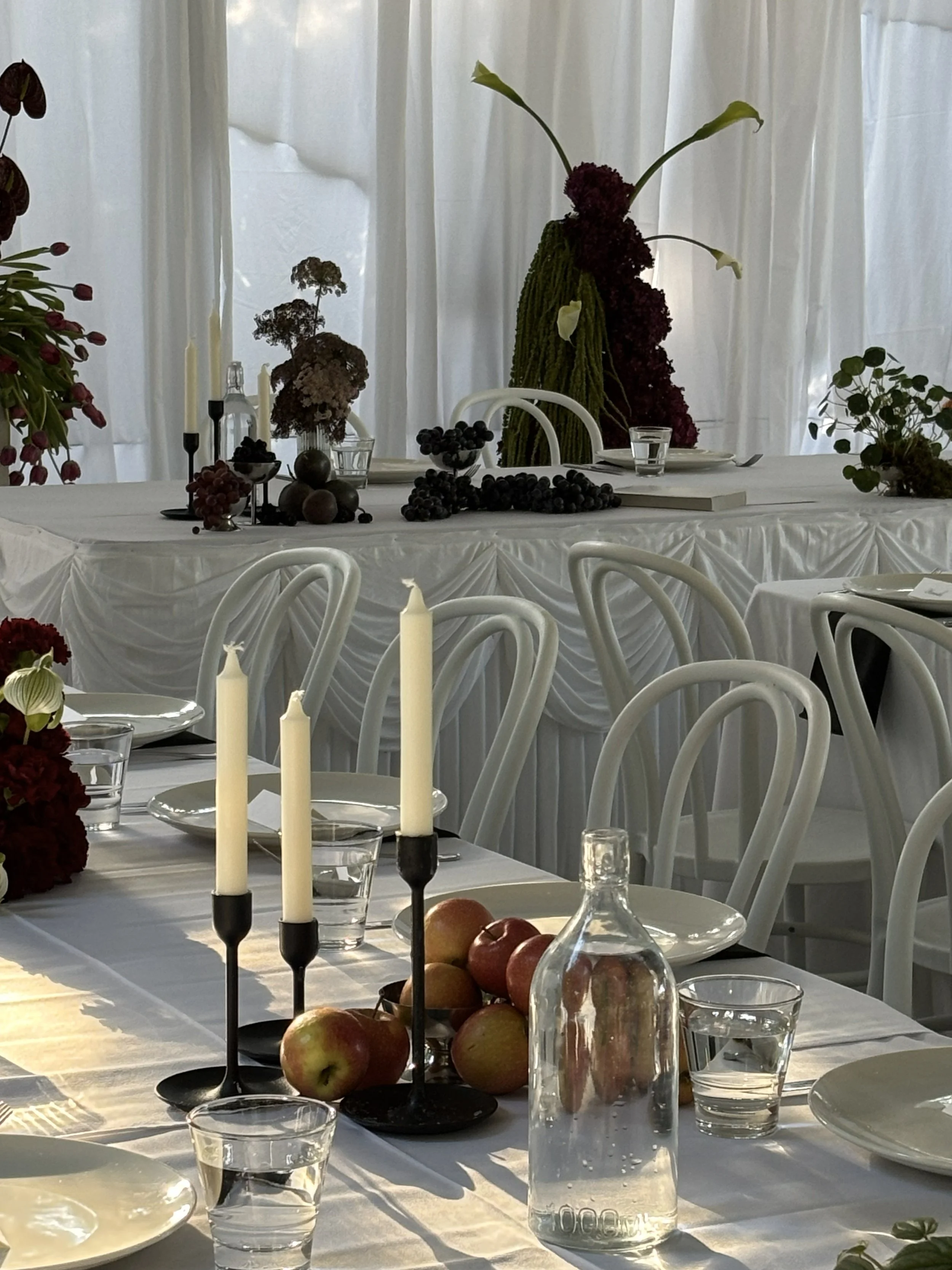 Elegant dining table setup with white chairs, white tablecloth, tall white candles in black holders, apples, water glasses, and floral centerpieces, with a decorated backdrop featuring flowers and draped white fabric.