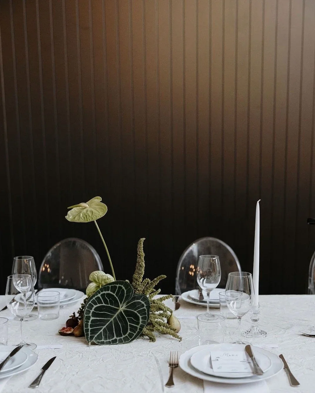 Elegant dining table set with white tablecloth, floral centerpiece with green leaves, clear glassware, white plates, silverware, and a single tall white candle against a dark wood-paneled wall.
