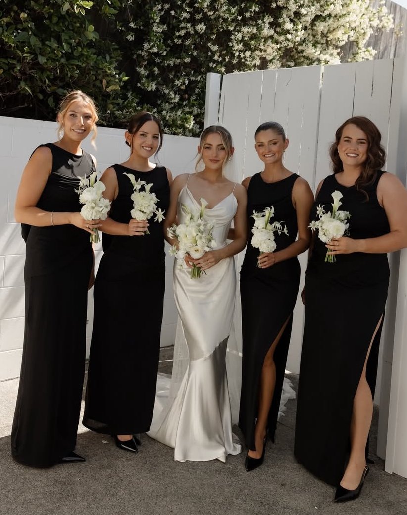 A bride in a white satin wedding dress holding a bouquet, surrounded by four bridesmaids in black dresses holding matching bouquets, standing outdoors against a white fence and greenery.