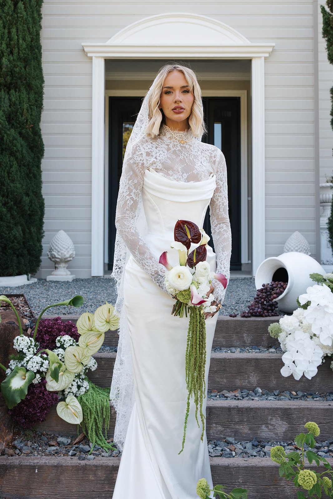 A woman in a white wedding dress with lace sleeves and high neckline standing on outdoor steps in front of a house, holding a bouquet of white and dark red flowers, surrounded by white and green floral decorations.