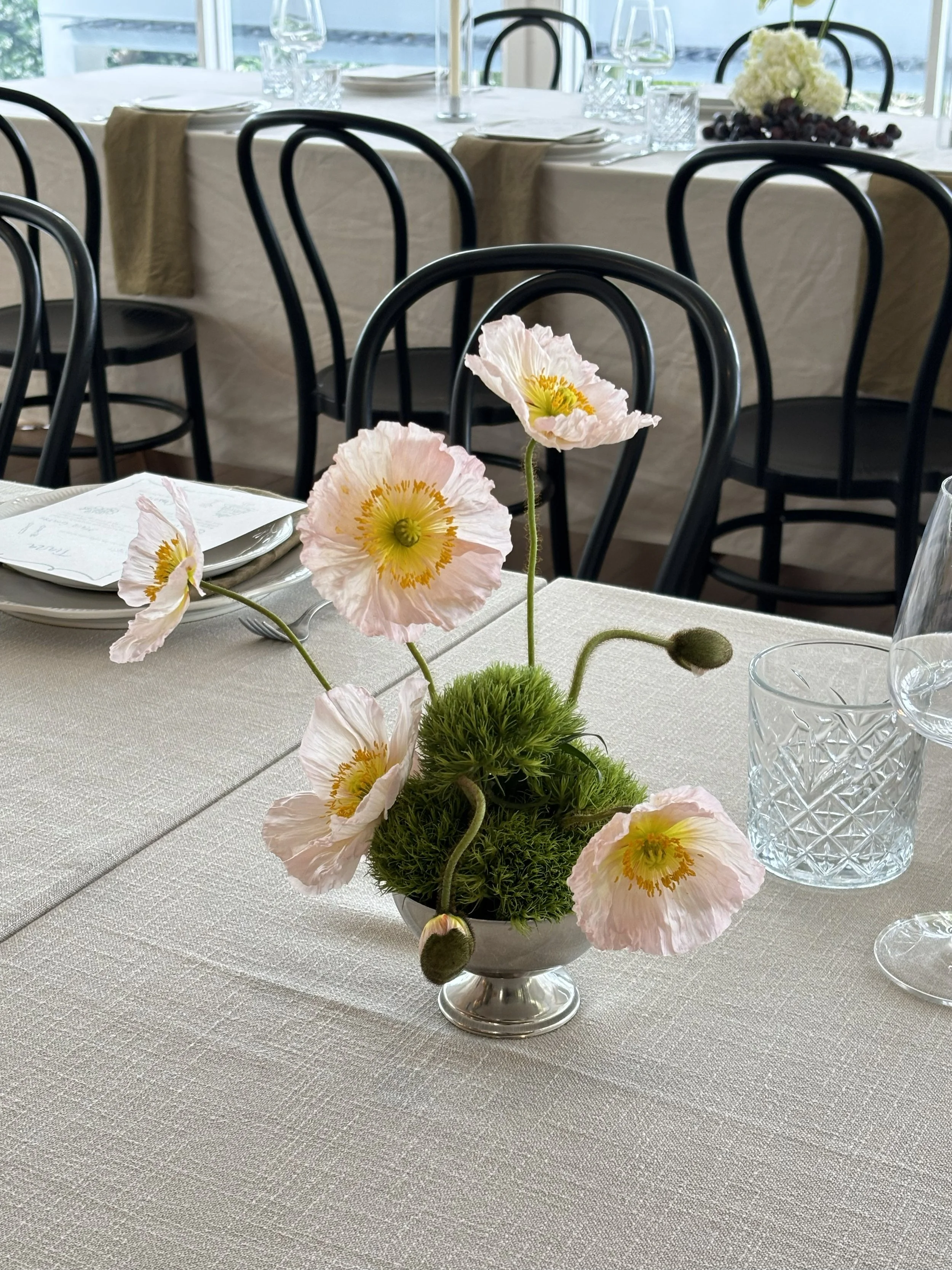 Elegant dining table with a centerpiece of pale pink poppy flowers and green moss in a silver vase, set with glassware, white napkins, and plates in a bright room.