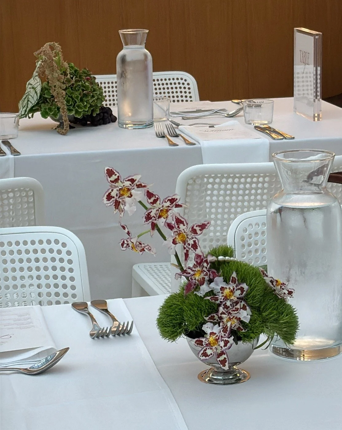 A table decorated with a white tablecloth, a floral arrangement with orchids and greenery, water pitchers, and place settings with utensils and glassware.