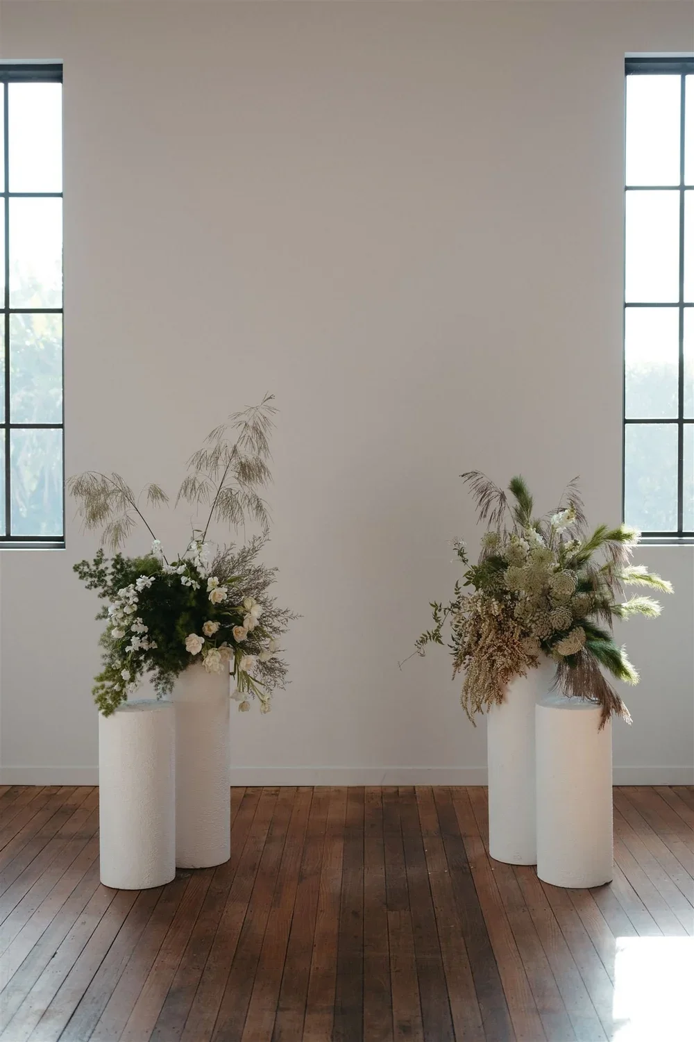 Two floral arrangements in white vases on white cylindrical pedestals against a plain white wall with large windows on each side, sunlight streaming in.