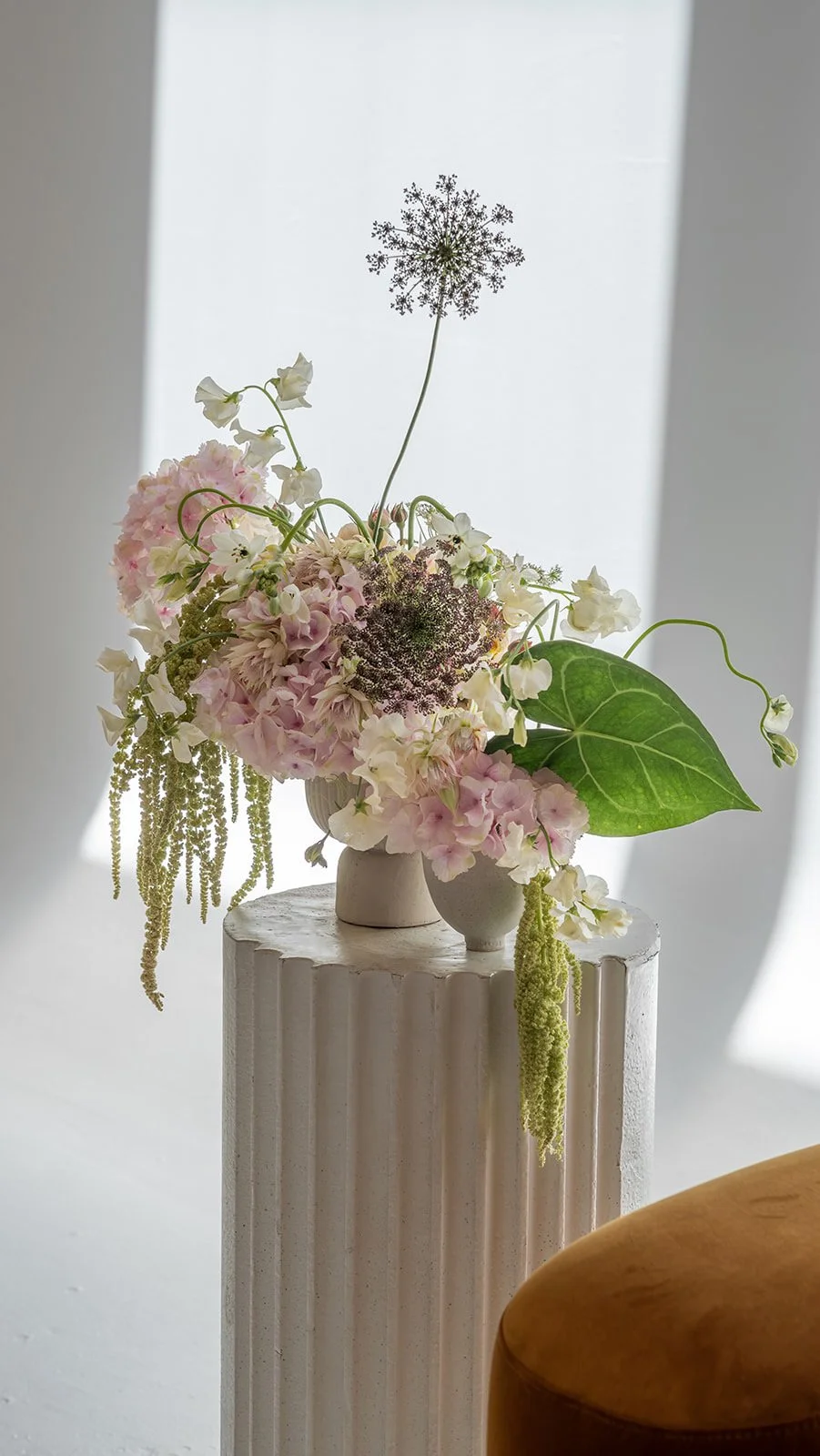A floral arrangement with pink and white flowers, green leaves, and tall seed heads in white vases on a white pedestal against a soft light background.
