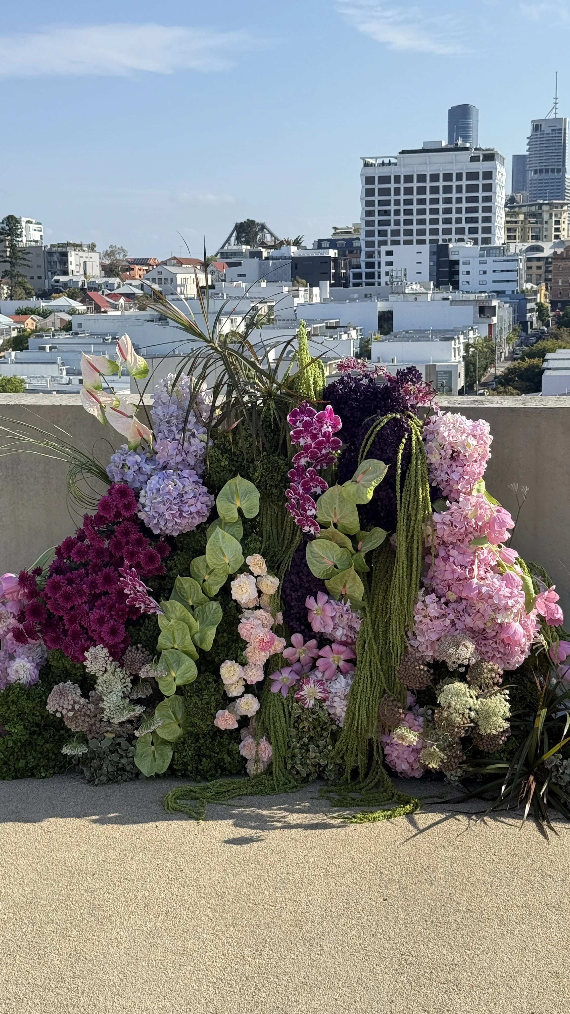 Colorful floral arrangement with pink, purple, and white flowers on a balcony with city skyline in the background.