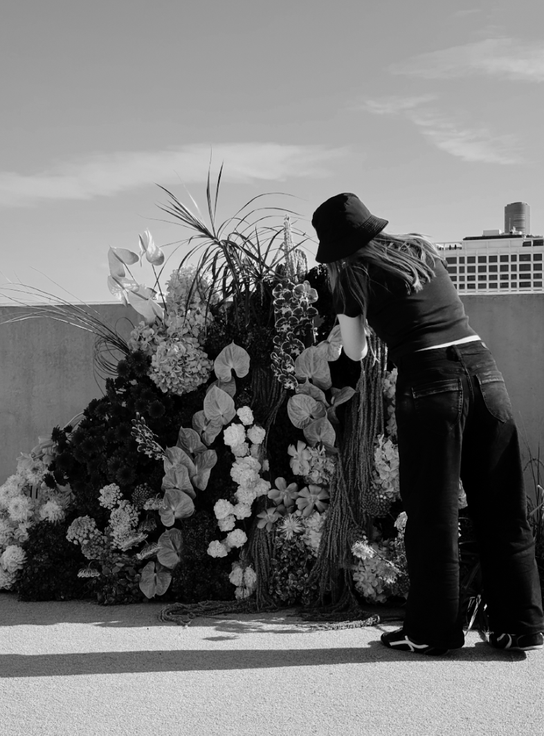 A woman with long hair, wearing a hat and casual clothes, arranging a large display of flowers on a rooftop terrace with a cityscape in the background.