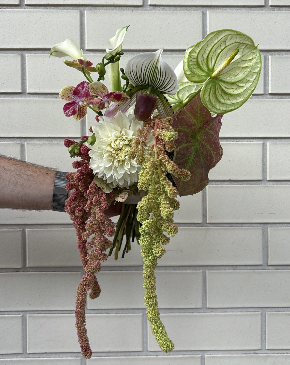 A hand holding a colorful bouquet of various flowers and greenery against a white brick wall.