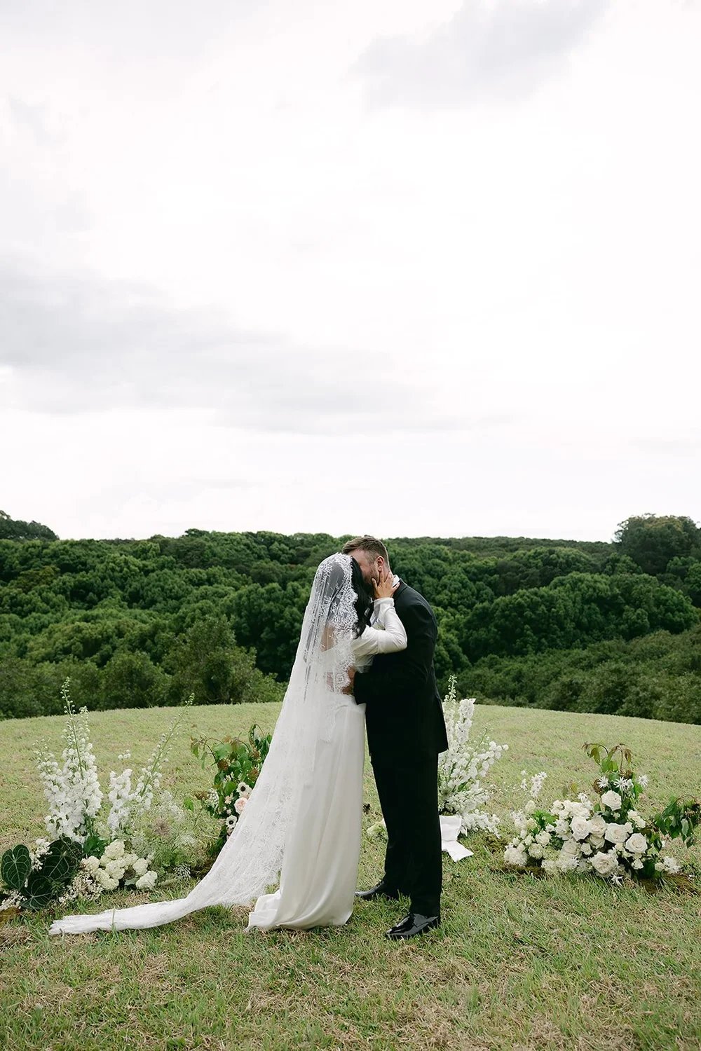 A bride and groom share a kiss outdoors on a grassy hill with a green wooded background and cloudy sky, surrounded by white floral arrangements.