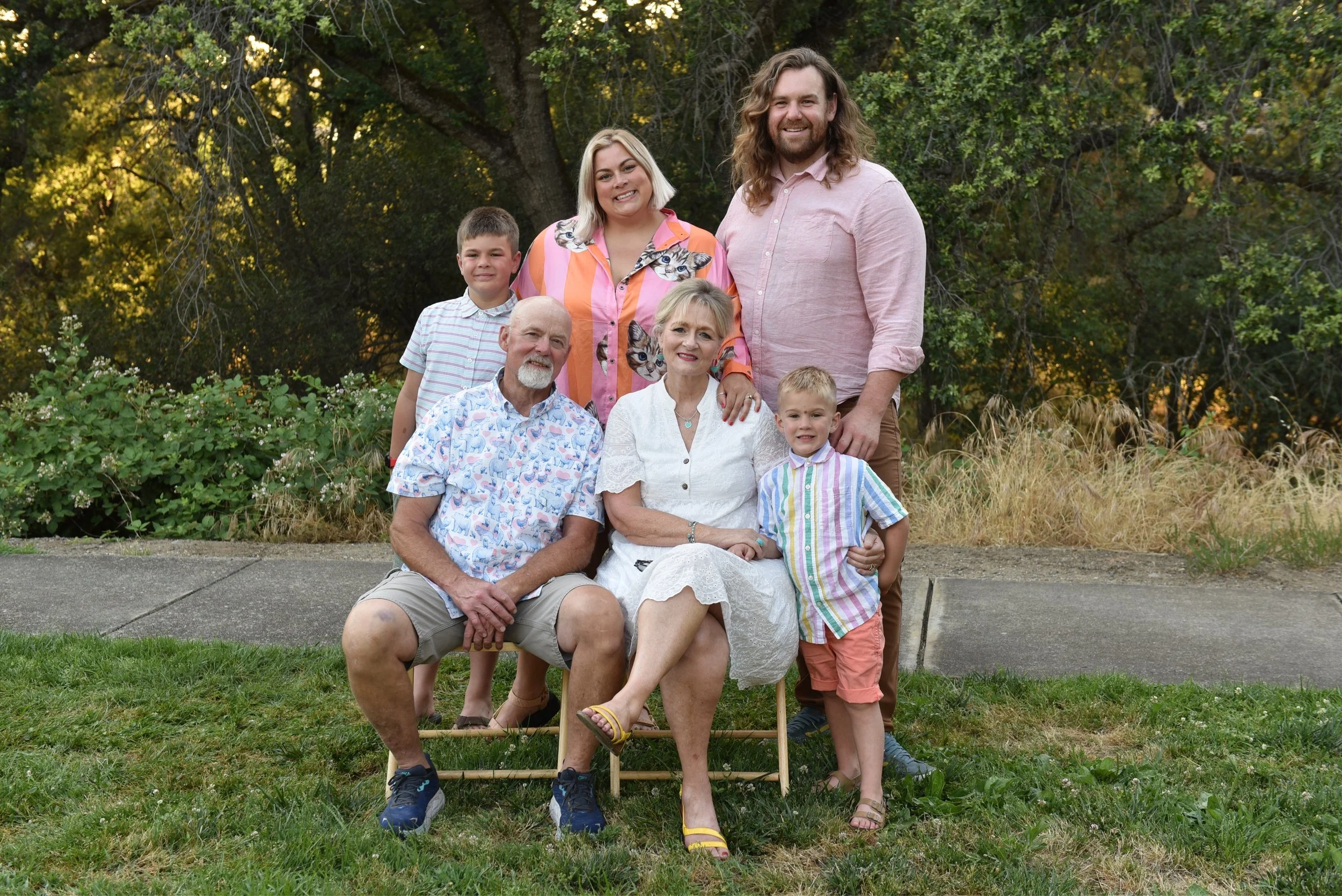 A multi-generational family portrait outdoors on a grass lawn, featuring three adults, two children, and an elderly woman, with trees and bushes in the background.