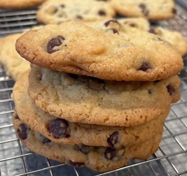 A stack of three chocolate chip cookies on a wire cooling rack, with more cookies in the background.