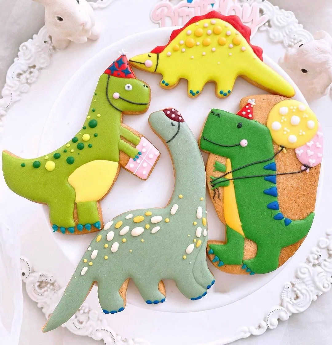Decorated cookies shaped like dinosaurs with colorful icing and sprinkles, arranged on a white plate with a lace border, surrounded by bunny figurines.
