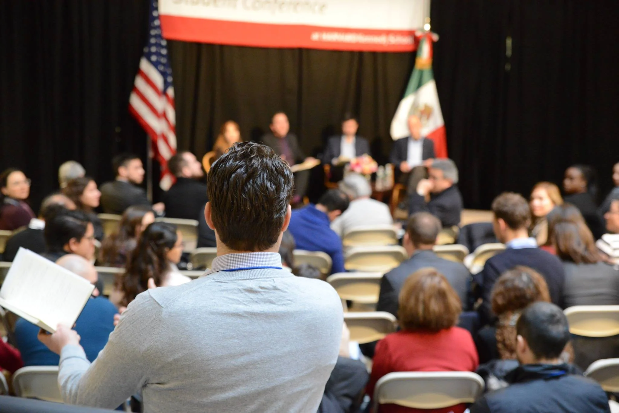 Reunión o conferencia con asistente escuchando atentamente, en el escenario hay panel de personas, banderas de Estados Unidos y México, en Harvard Kennedy School.