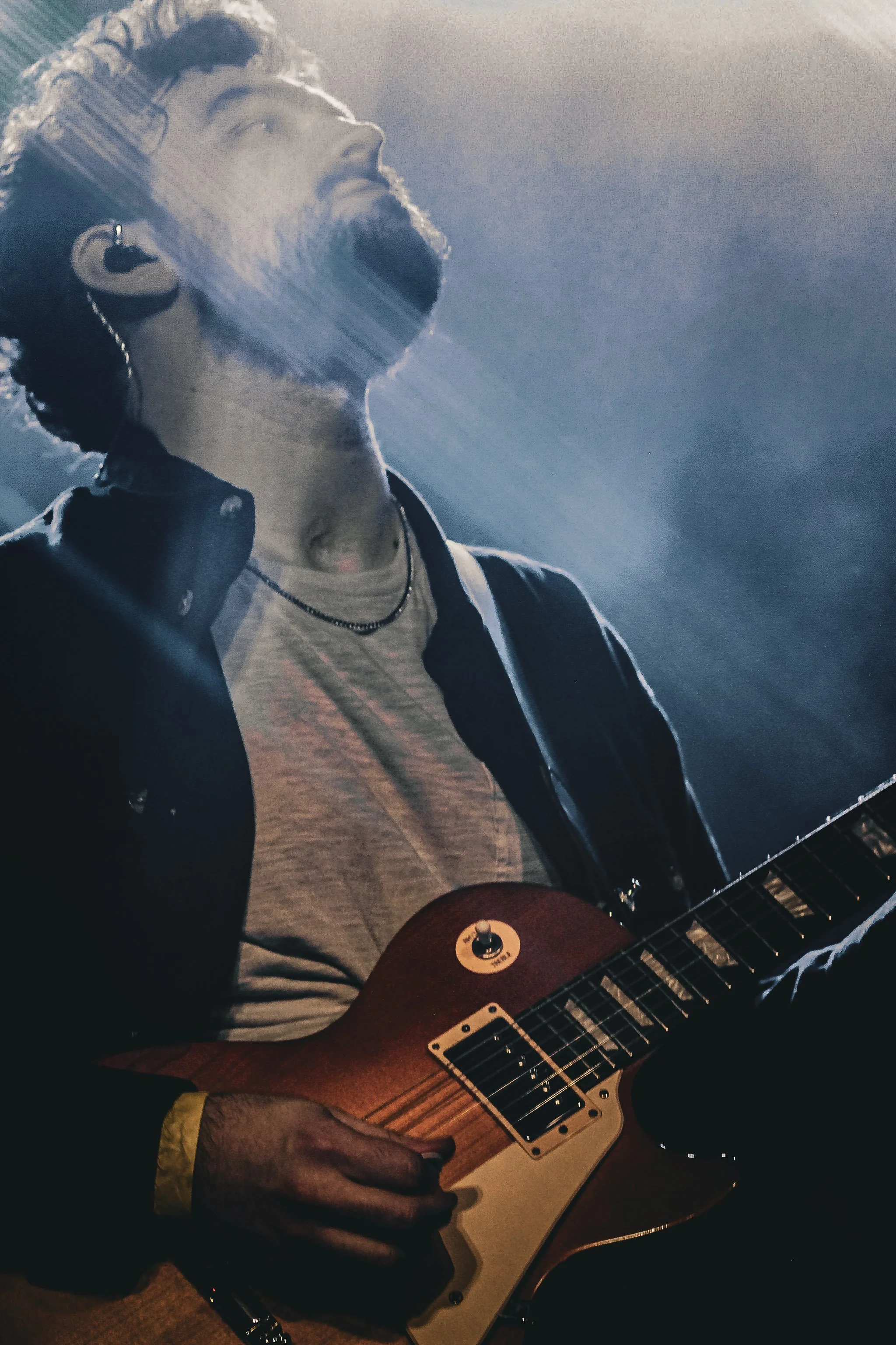 A musician playing an electric guitar on stage with dramatic lighting, wearing a grey t-shirt, dark jacket, and jewelry.