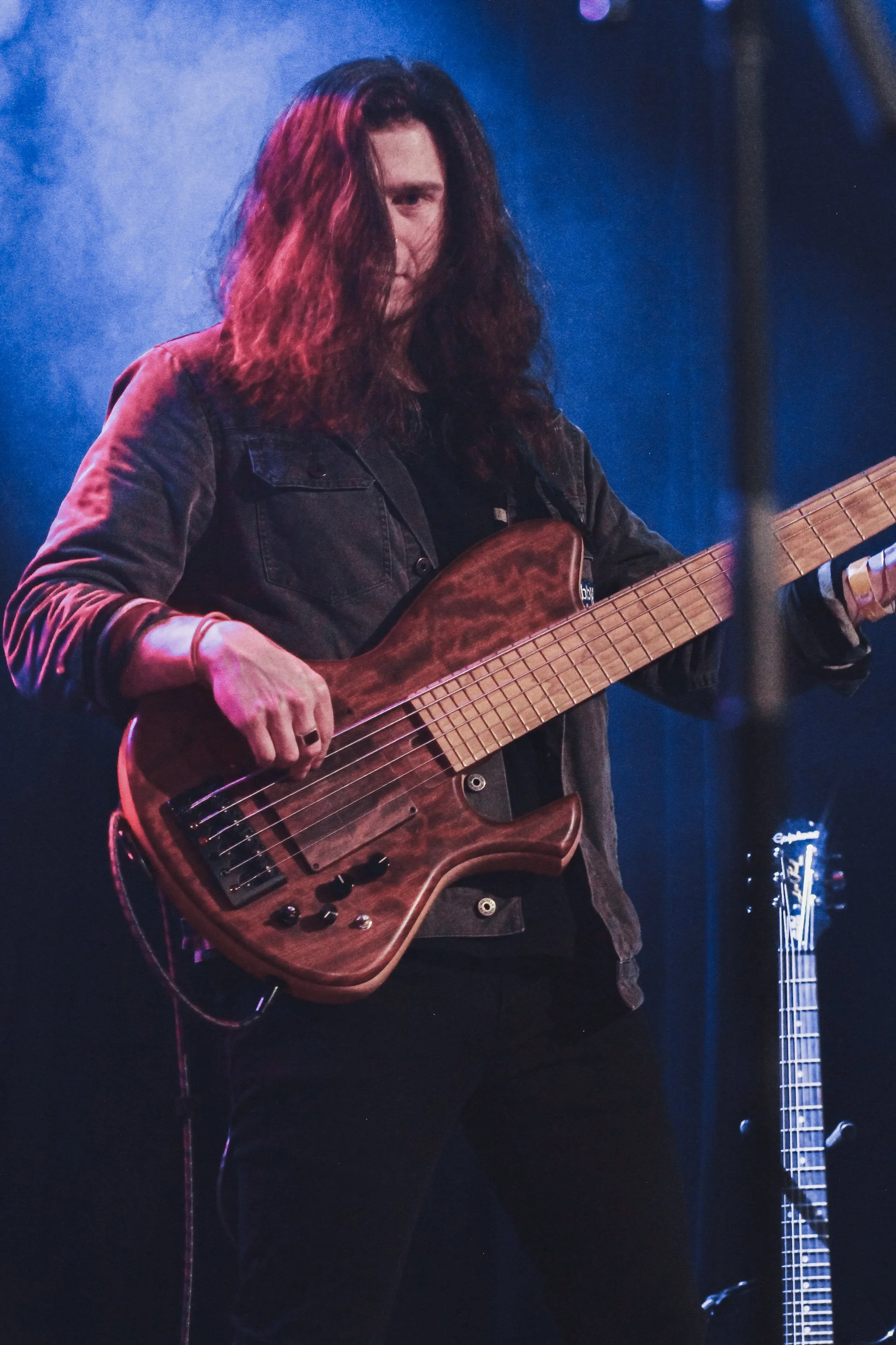 A musician with long curly hair, wearing a dark jacket, playing a wooden bass guitar on stage with blue lighting.