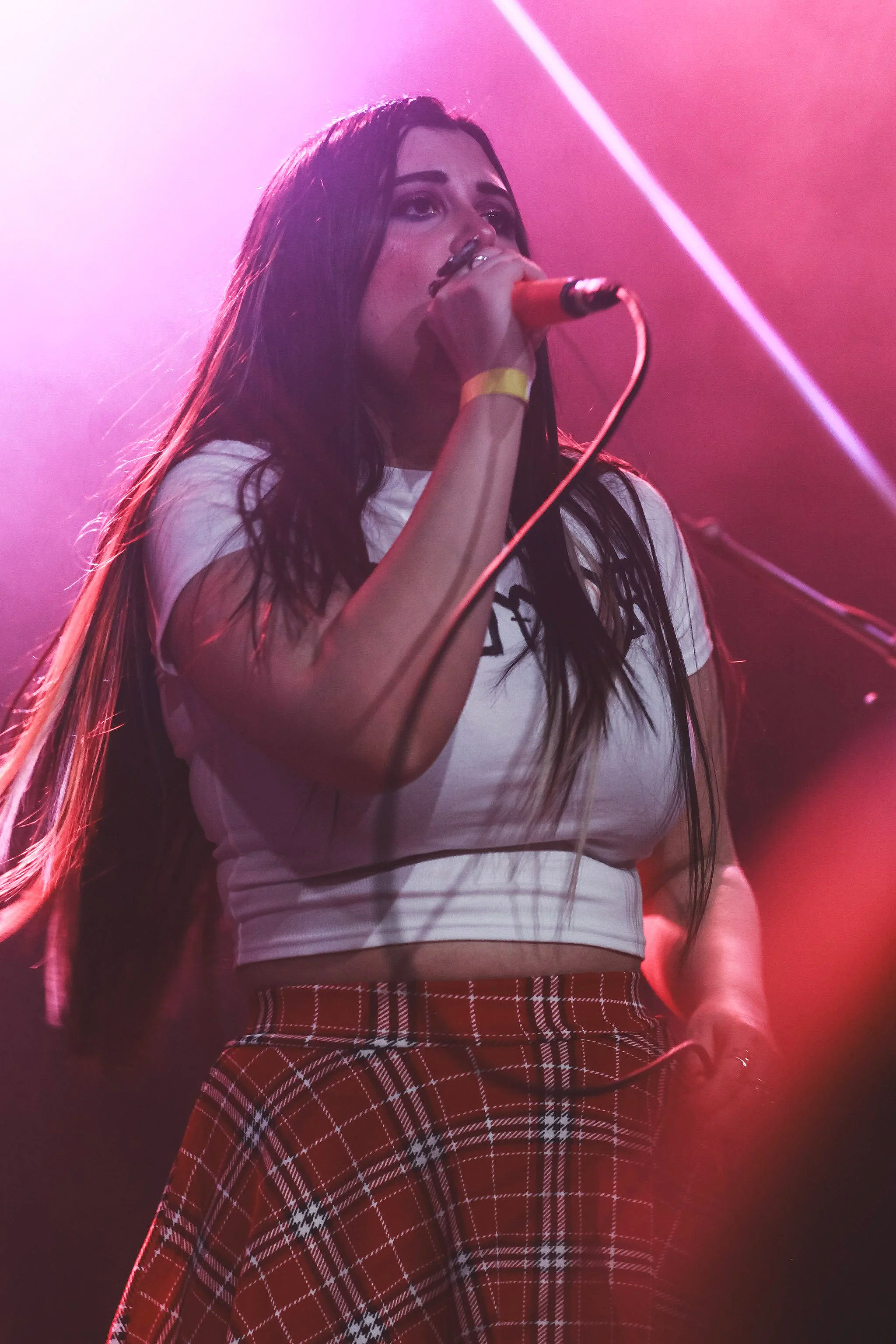 A woman with long dark hair singing into a microphone on stage under pink lighting.
