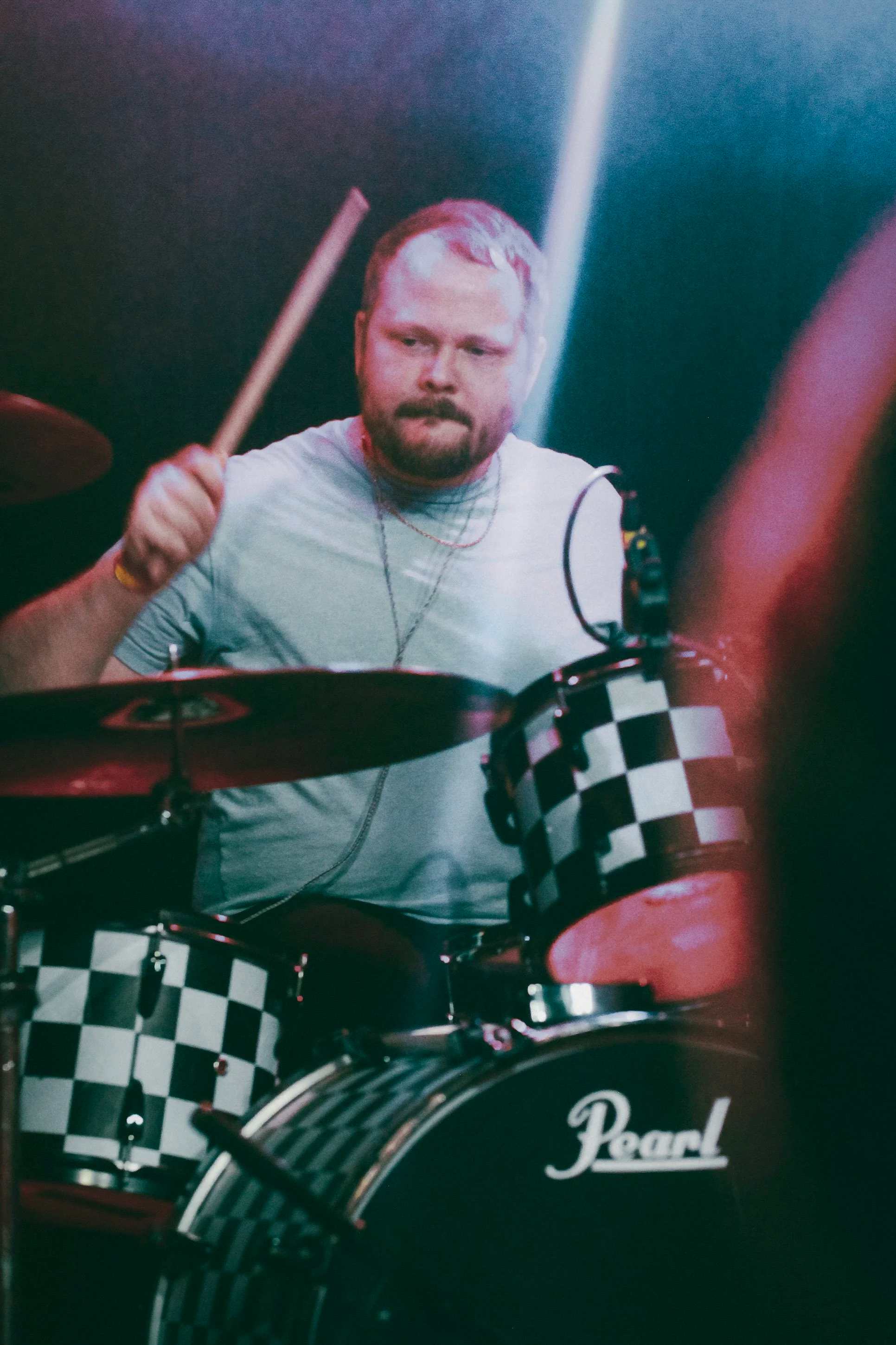 A man with a beard playing a drum set, wearing a light grey t-shirt and headphones, with a black and white checkered drum kit with 'Pearl' branding.