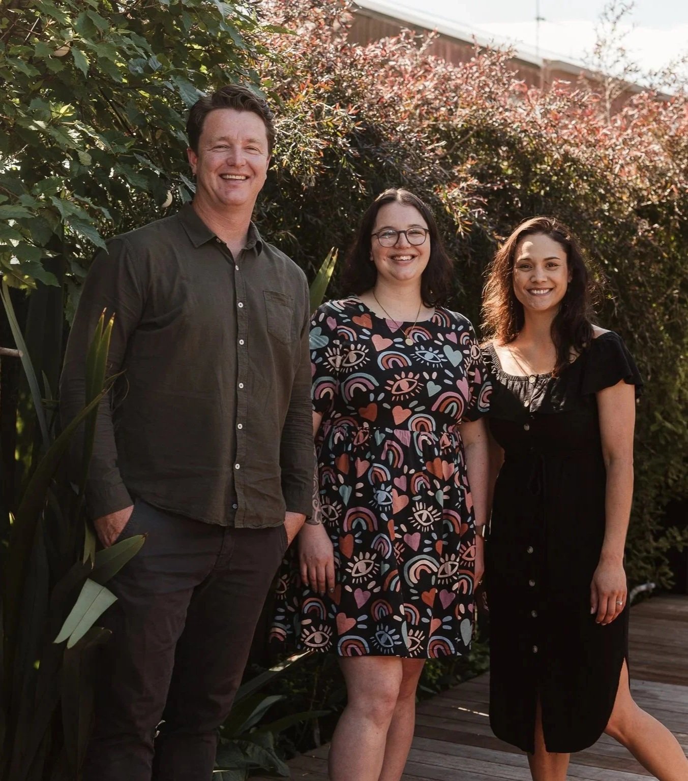 Three people standing outdoors in a garden with plants and trees, smiling at the camera.