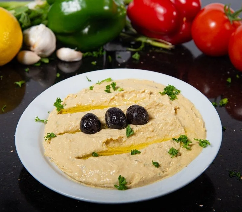 A white plate of hummus garnished with herbs and olives, with tomatoes, garlic, and green bell pepper in the background.