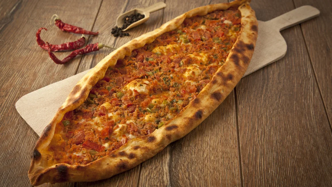 A large, boat-shaped pizza on a wooden table with chili peppers and peppercorns nearby.