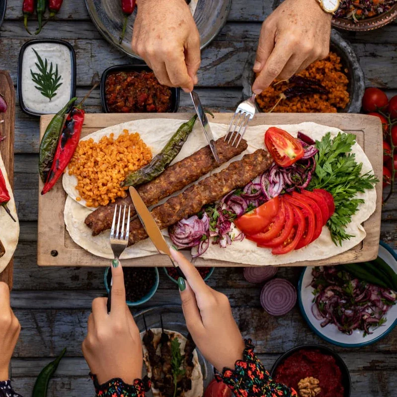 A wooden table with a platter of grilled kebabs, slices of tomatoes and red onions, fresh parsley, and rice, surrounded by salads and condiments, with hands reaching for food.