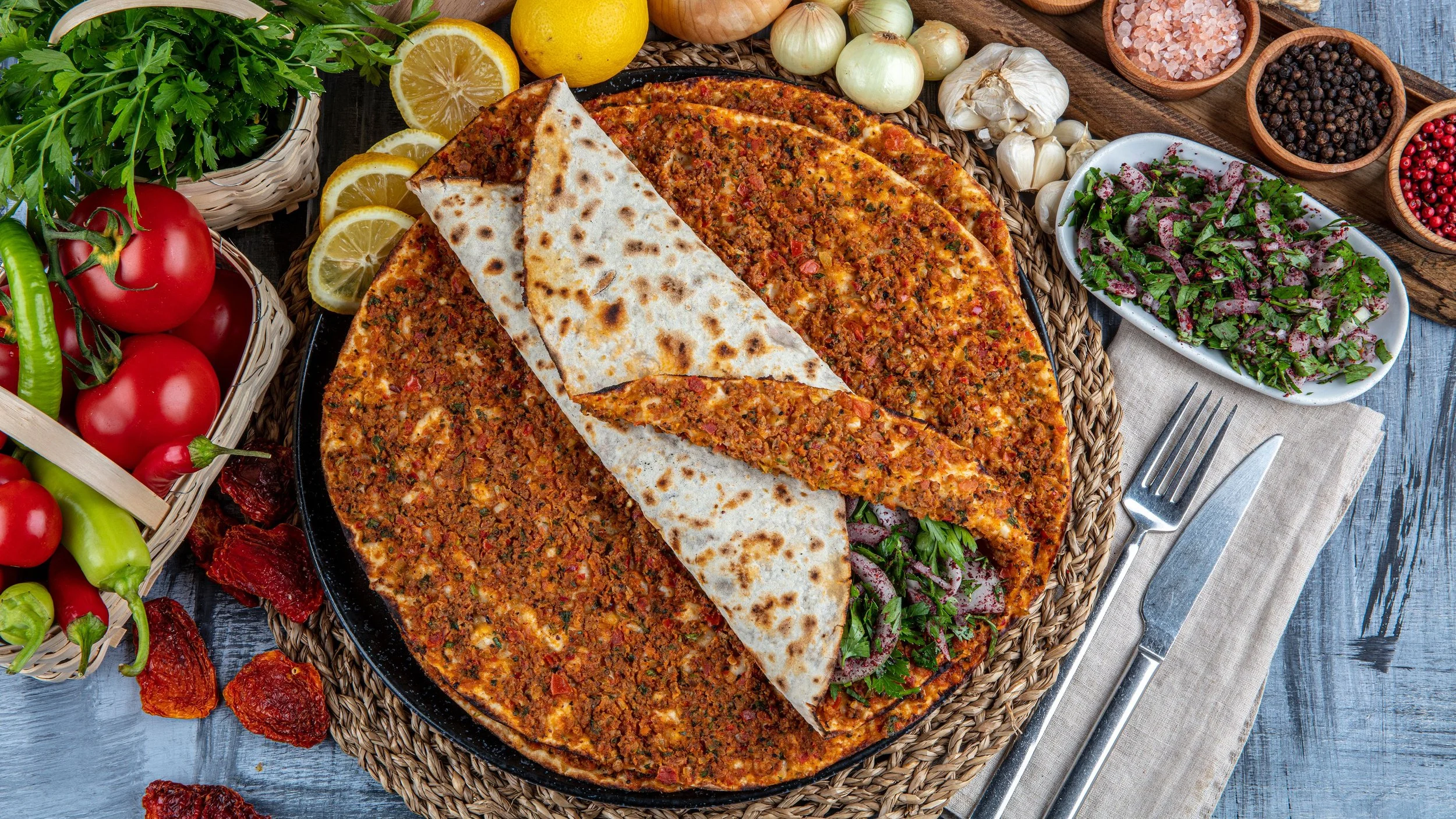 A Turkish-style Lahmacun or flatbread with minced meat and vegetables, served with lemon slices, surrounded by fresh vegetables and herbs, garlic, onions, spices, and a green salad on a rustic wooden table.