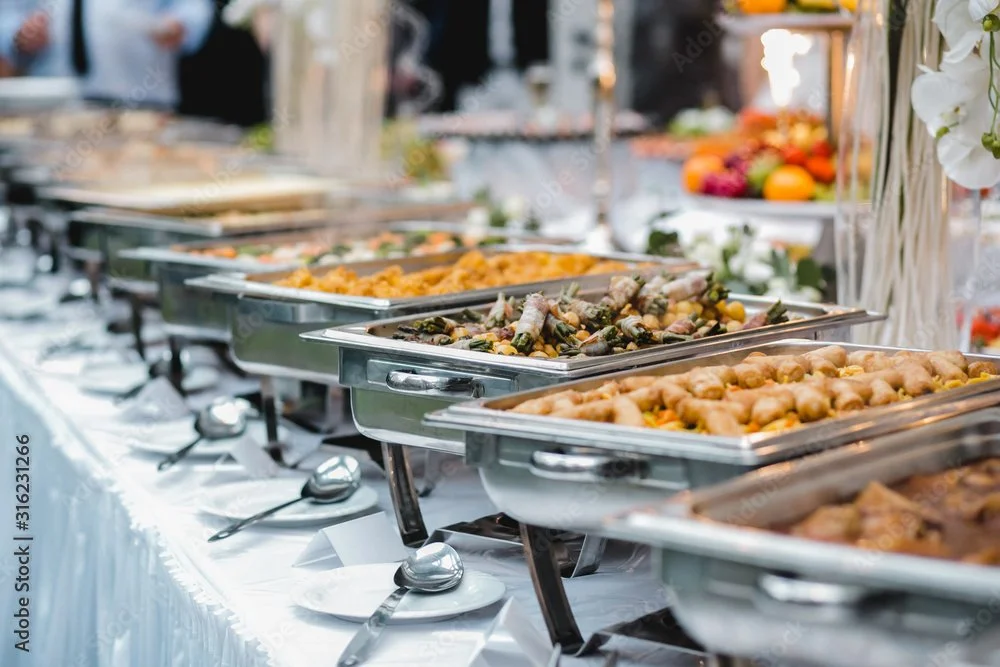 Buffet table with stainless steel chafing dishes filled with various hot foods, set outdoors at a sunny event with white tablecloths and floral decorations.