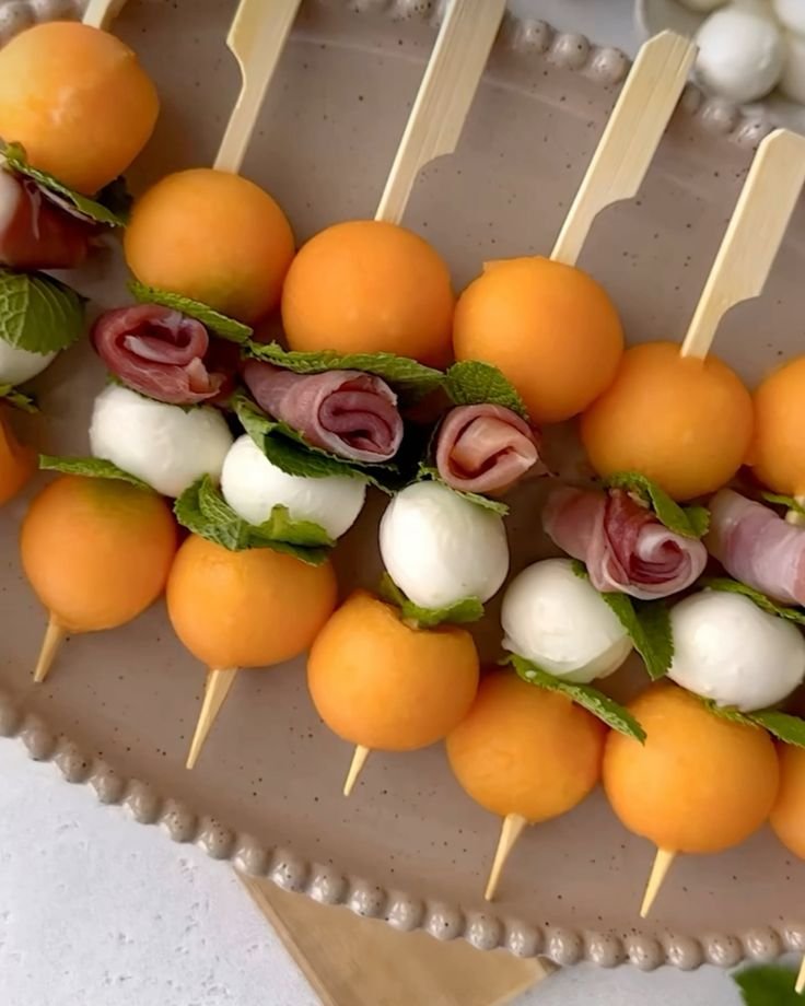 Skewers with orange melon balls, mozzarella balls, prosciutto, and mint leaves on a platter.