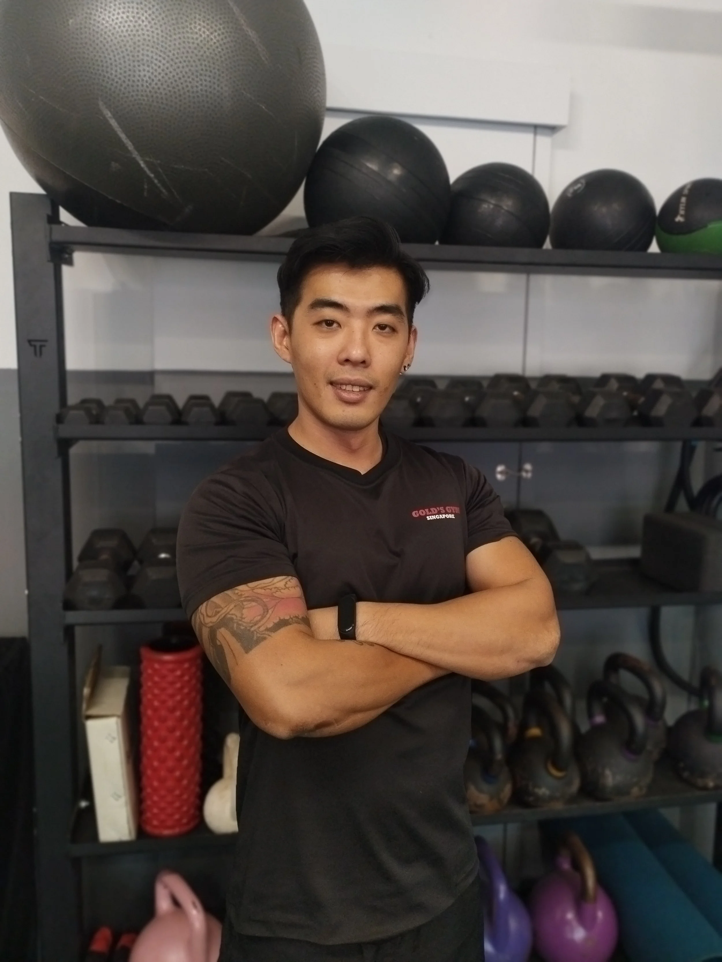 A young man with black hair and a tattoo on his right arm standing in a gym with arms crossed, black workout shirt, in front of shelves with kettlebells and medicine balls.