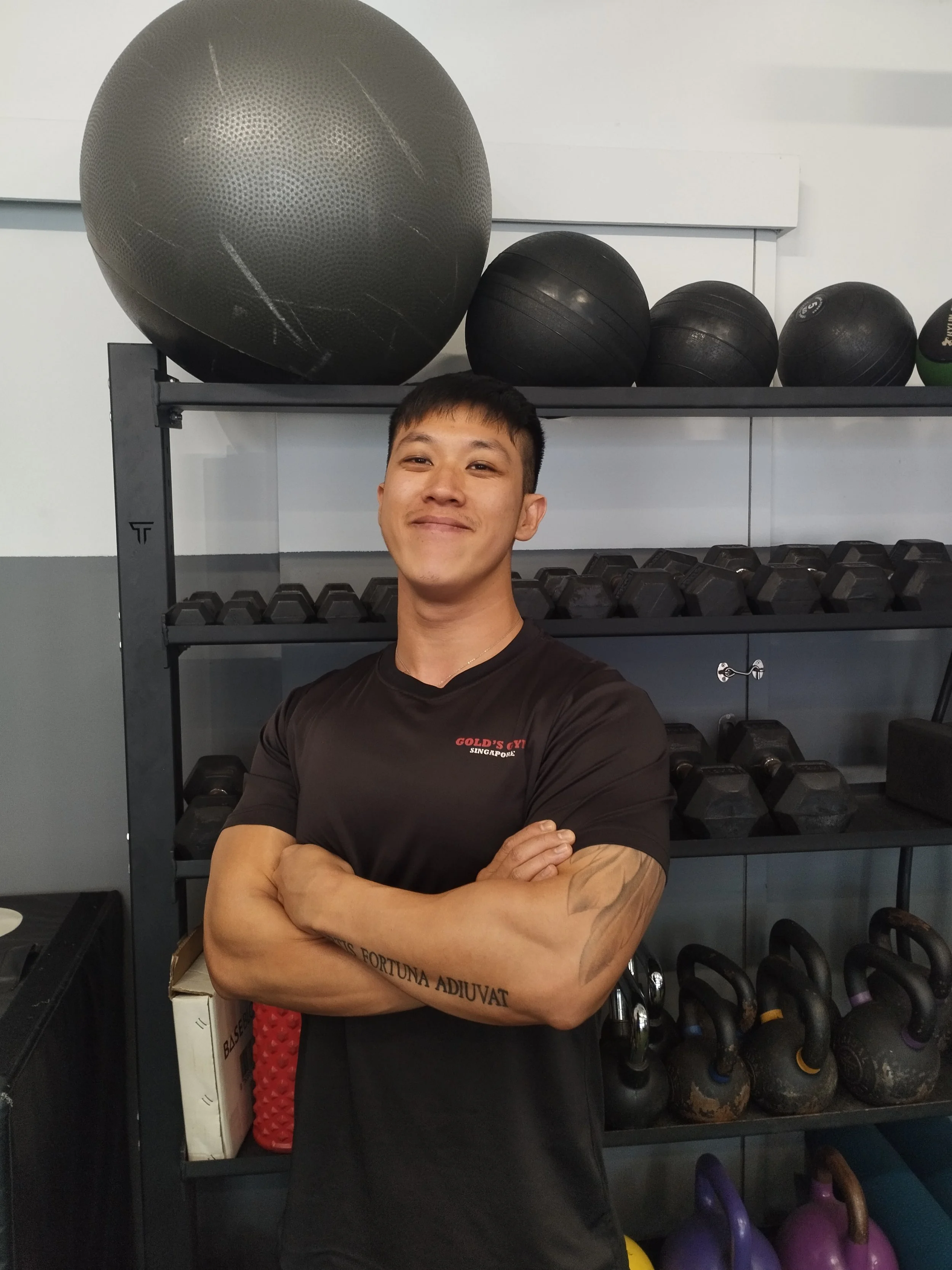 Young man with crossed arms smiling in gym with black weights and exercise balls on shelf behind him.