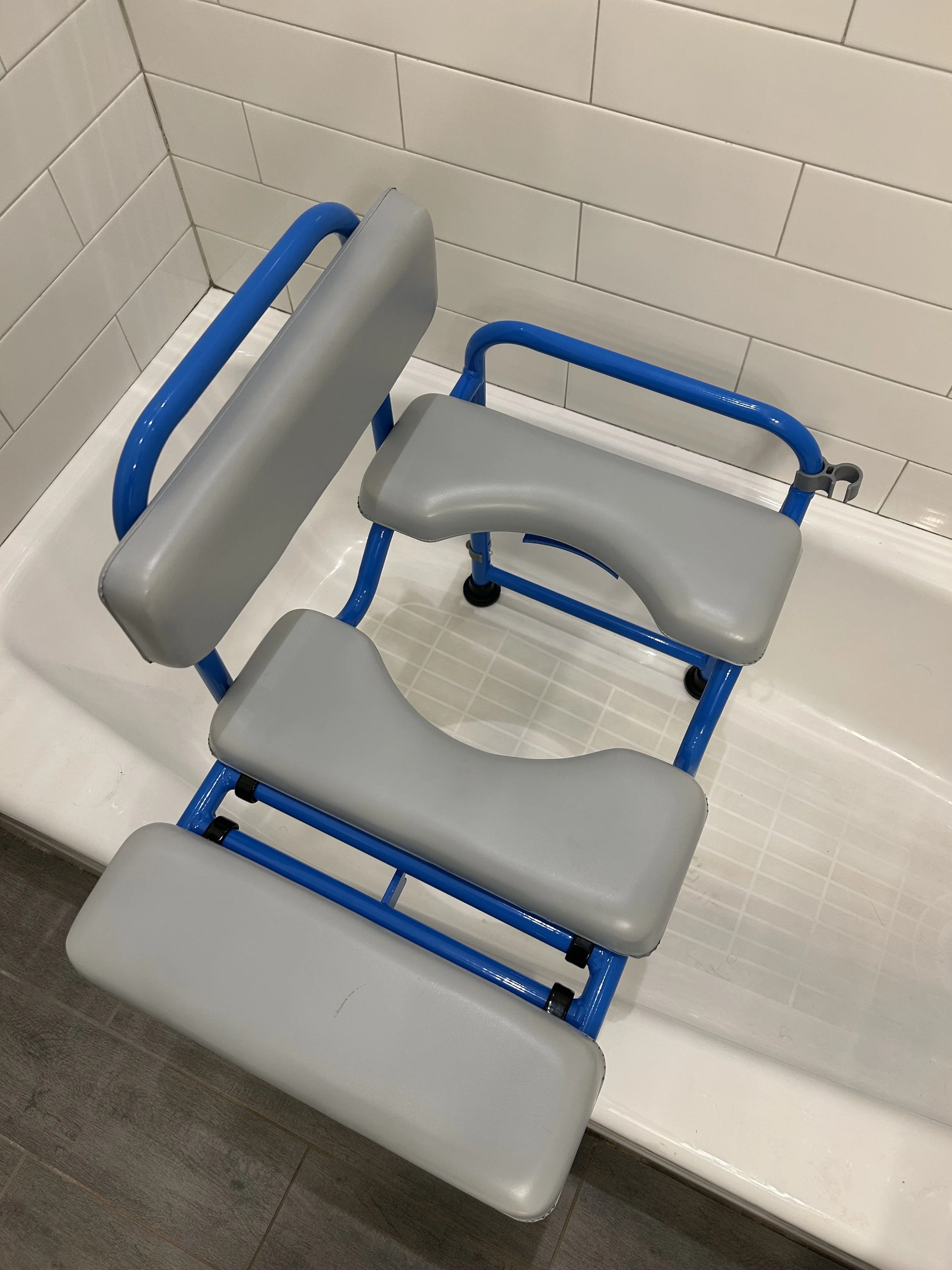 A shower chair with gray cushioned seats and blue frame, situated in a shower with white tiled walls.