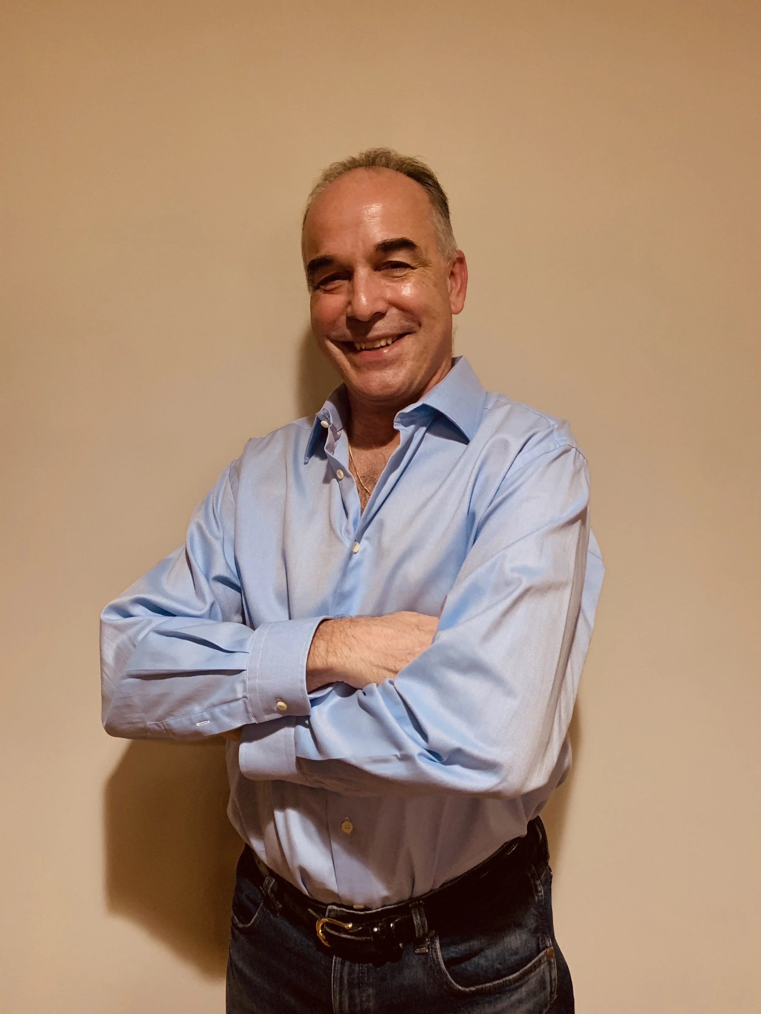 Smiling man with crossed arms wearing a light blue dress shirt and dark jeans, standing against a plain beige wall.