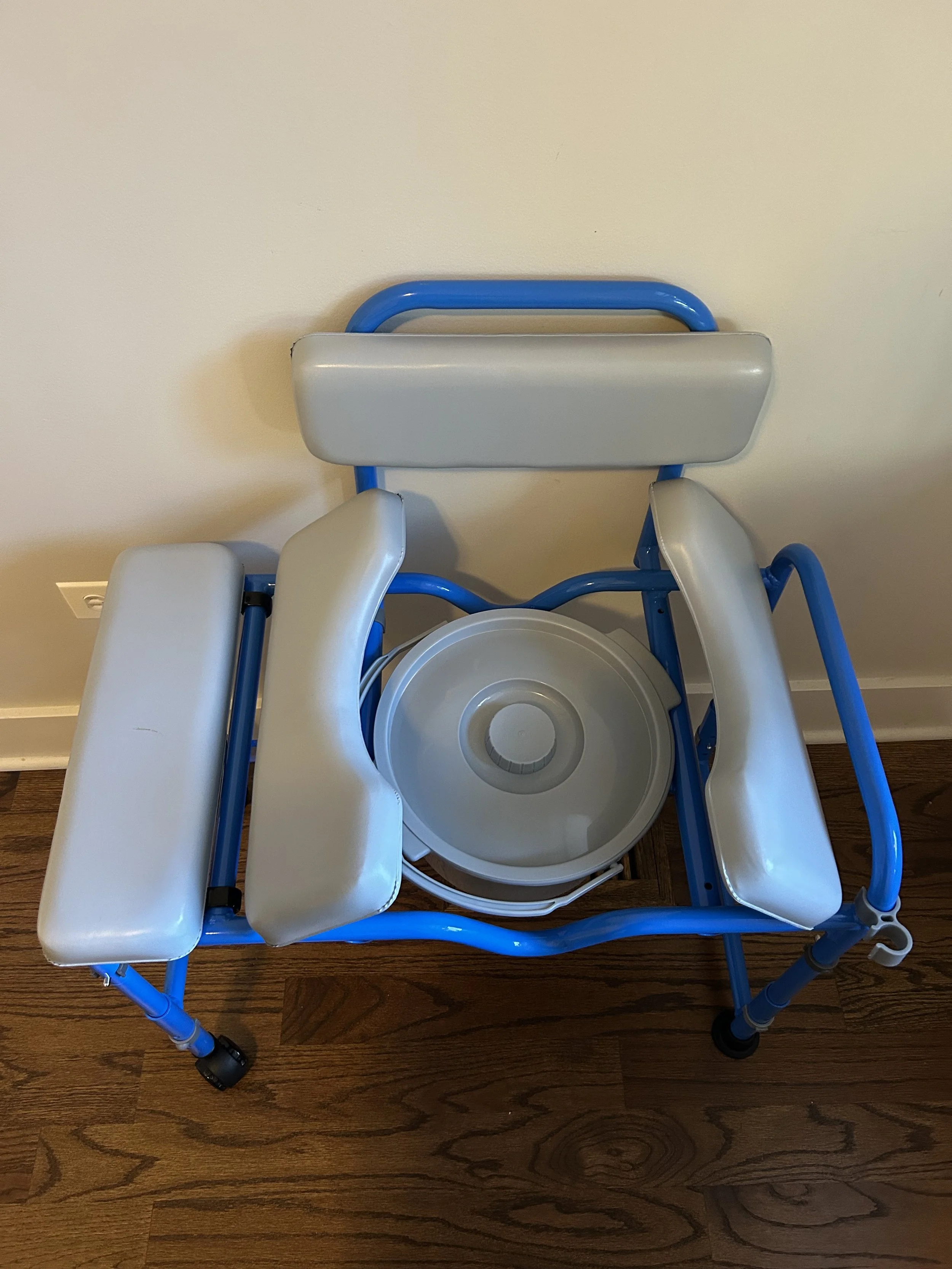 A blue and white shower chair with armrests and a circular footrest, positioned on a wooden floor against a plain wall.