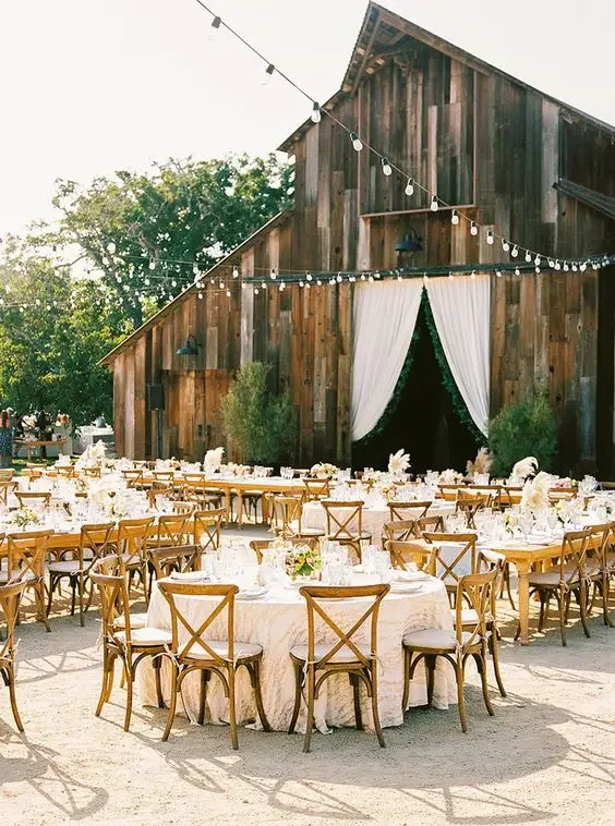 an-elegant-outdoor-barn-wedding-reception-space-with-round-tables-pastel-blooms-and-greenery-pampas-grass-and-string-lights-over-the-space.webp